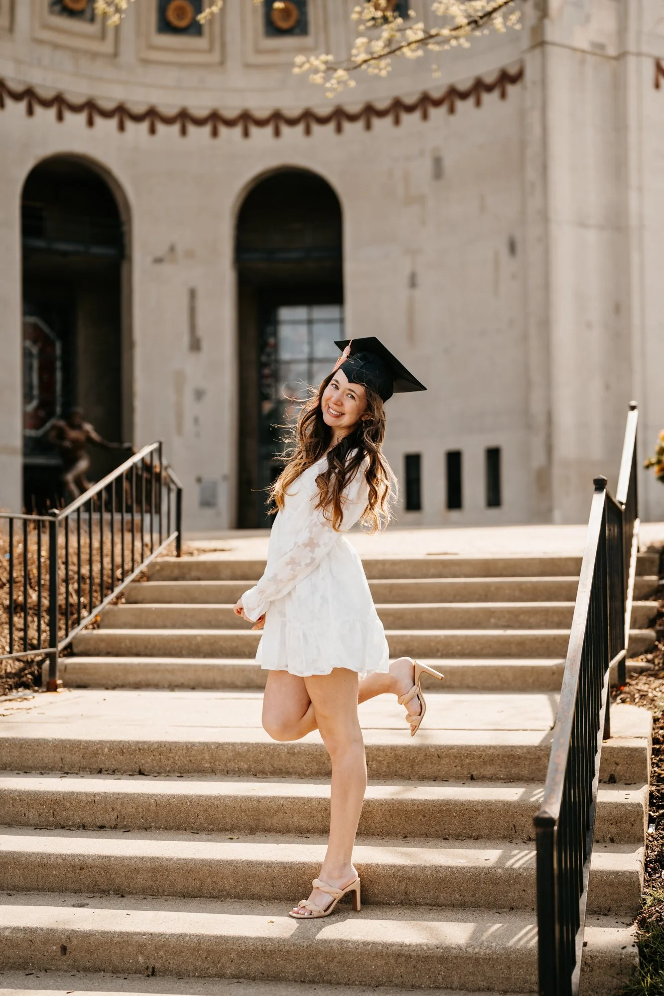 A young woman in a white dress and high heels, wearing a graduation cap, standing on outdoor stairs and posing with one foot lifted, smiling at the camera.