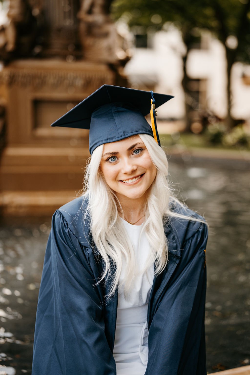 A young woman with long blonde hair and blue eyes dressed in a dark graduation gown and cap, smiling outdoors.