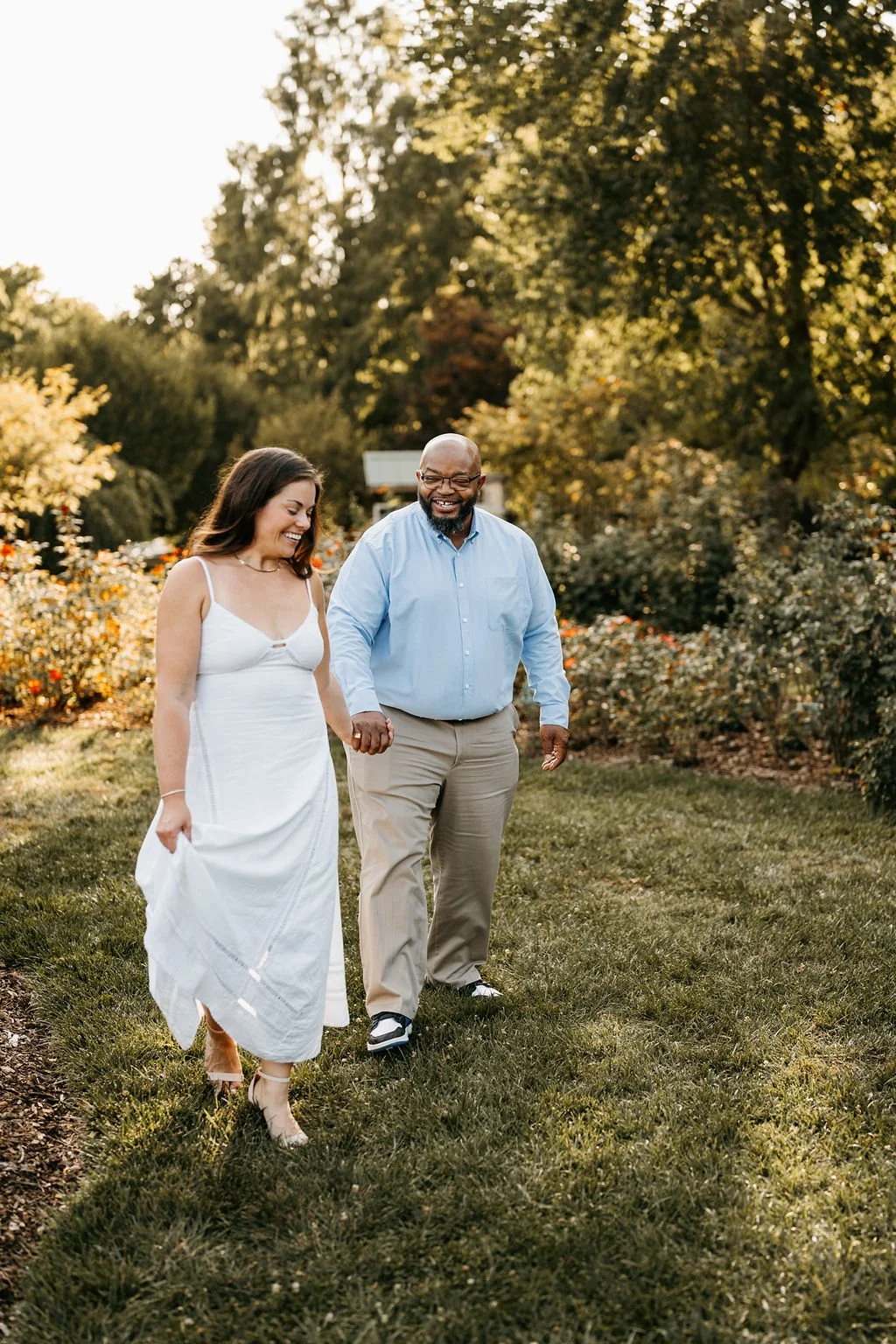 A couple walking hand in hand outdoors in a garden or park during sunset, smiling and enjoying each other's company.