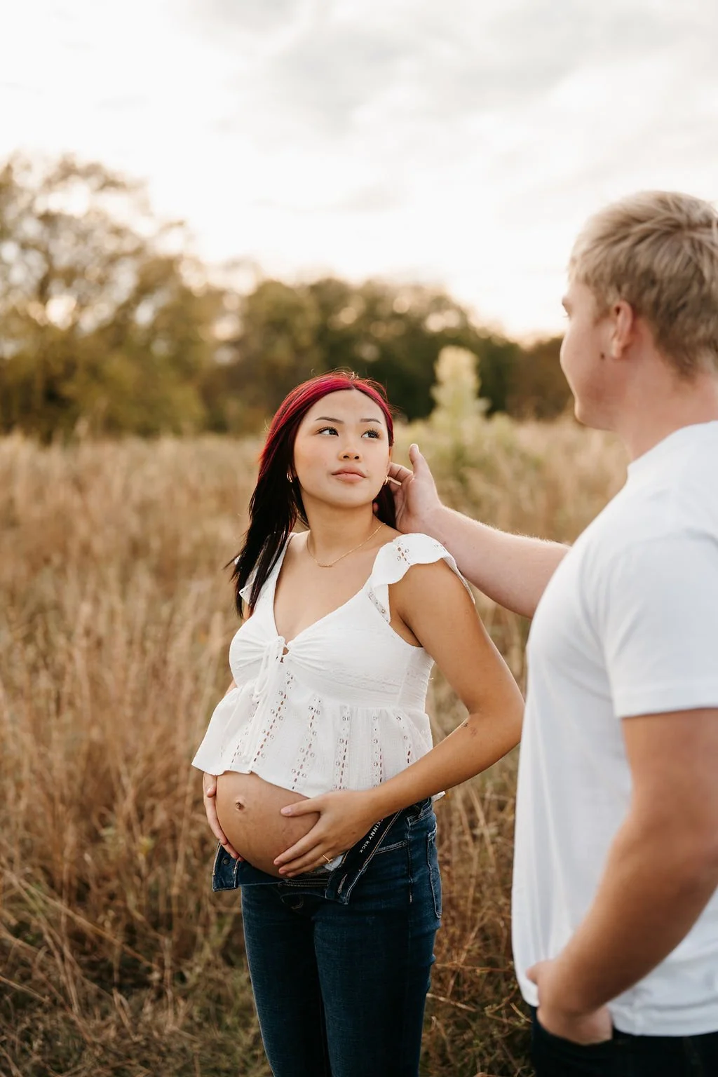 Pregnant woman in a white top and jeans standing in a field, with a man touching her face, in an outdoor setting during sunset.