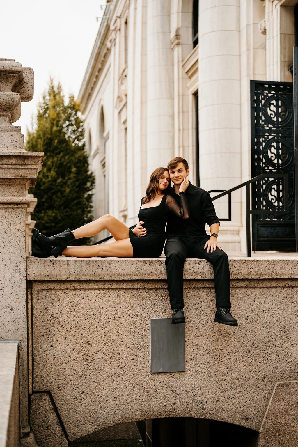 A young couple sitting on a stone ledge outside a historic building, with the woman leaning on the man's shoulder, both dressed in black outfits, with trees and ornate architecture in the background.