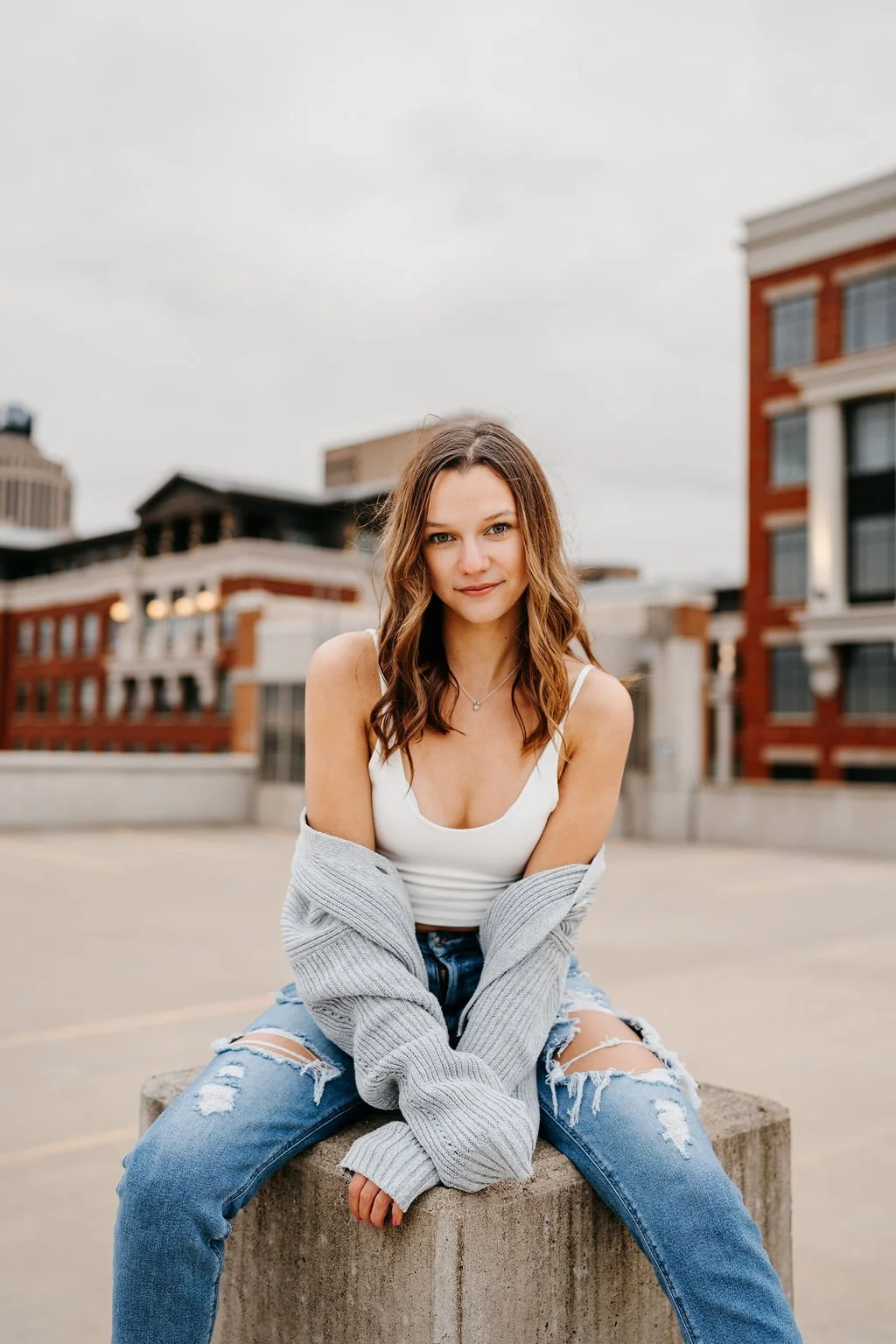 A young woman with brown hair and a white tank top sitting on a concrete block outdoors, with urban buildings in the background.