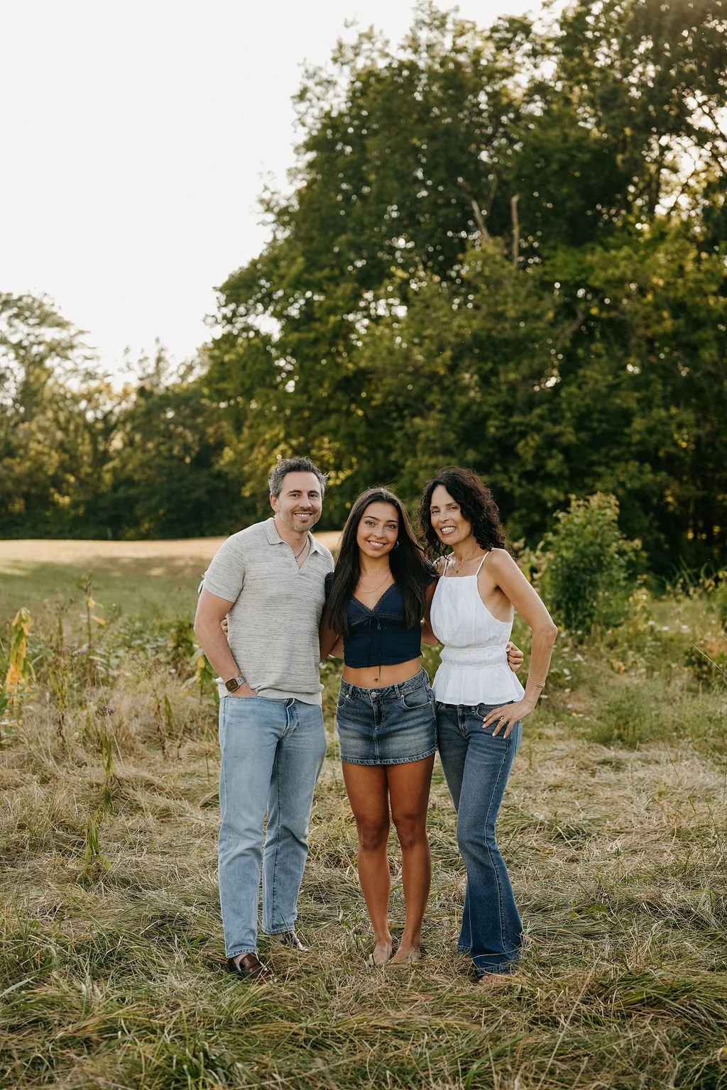 Three friends standing outdoors in a grassy field with trees in the background, smiling at the camera.