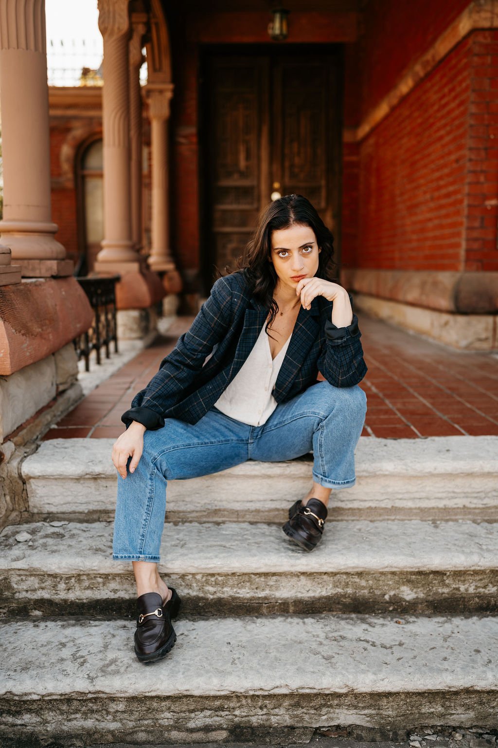 Young woman with dark hair, dressed in a plaid blazer, white blouse, and jeans, sitting on outdoor steps with a brick building in the background.