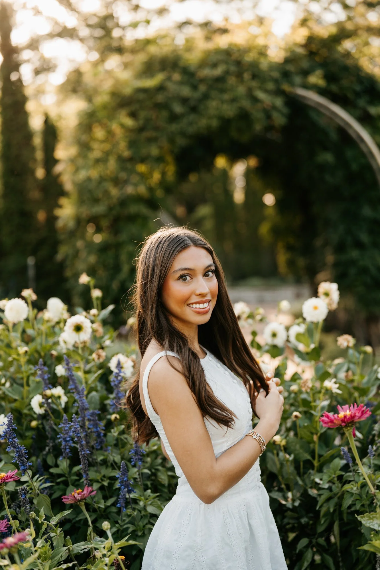 A woman standing in a garden of flowers during sunset, smiling at the camera.