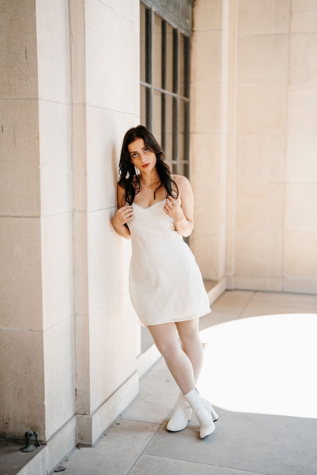 Young woman with dark wavy hair and fair skin, wearing a white slip dress and white ankle boots, standing against a light-colored stone wall near a glass window, with one hand holding a strap of her dress and the other near her shoulder.