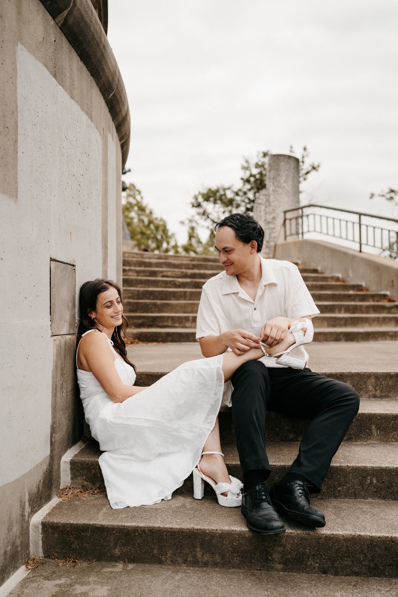 A woman in a white dress sitting on stairs, leaning against a wall, smiling at a man in a white shirt and black pants, who is feeding her a baby while sitting on stairs outdoors.
