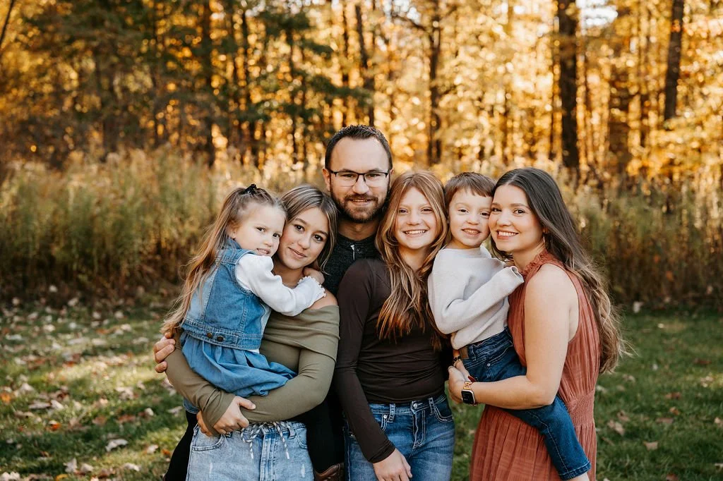 Family of six smiling and embracing outdoors during fall, with trees and autumn leaves in the background.