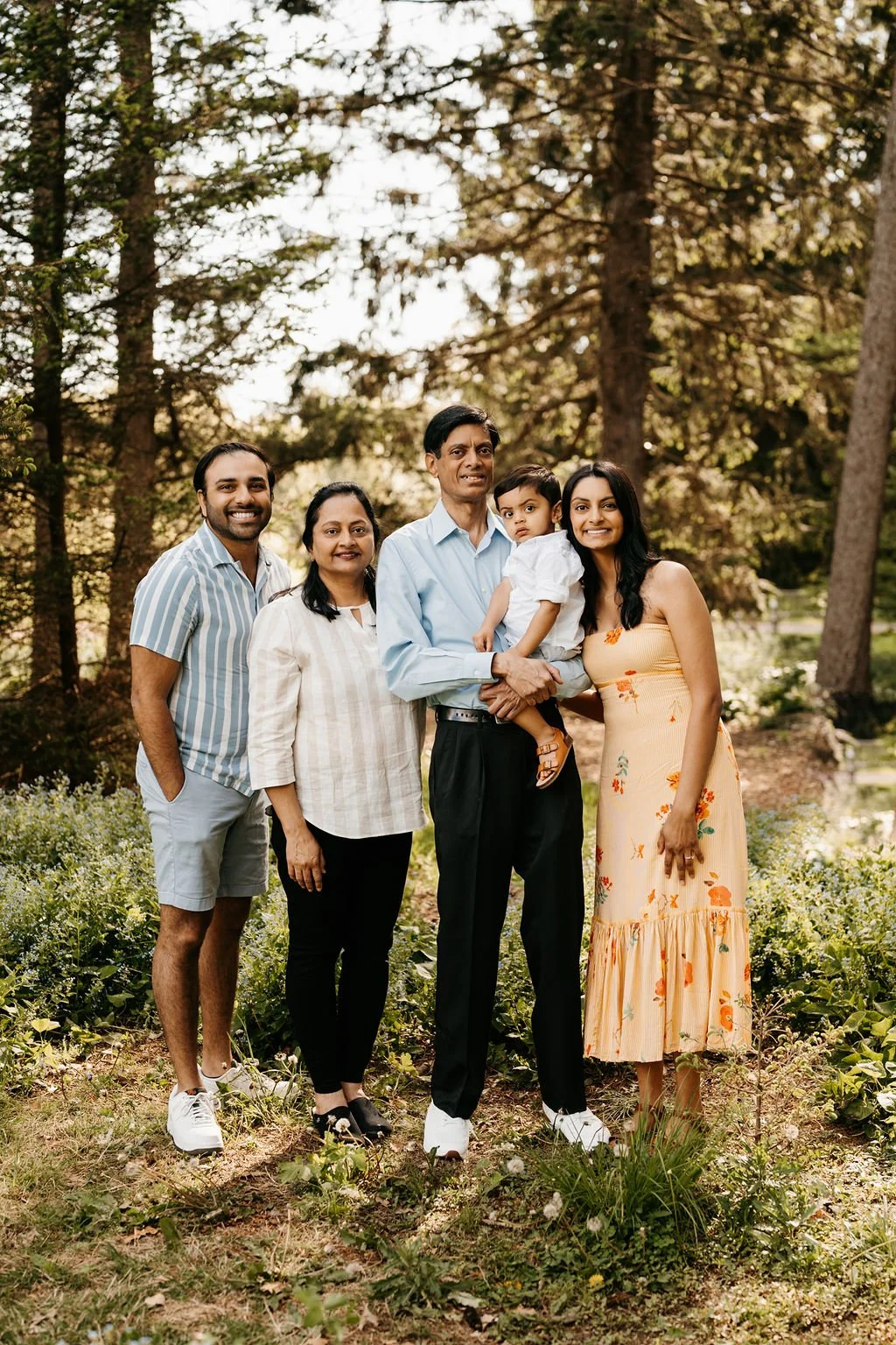 Family of five standing outdoors on a sunny day in a wooded area, smiling for a group photo.
