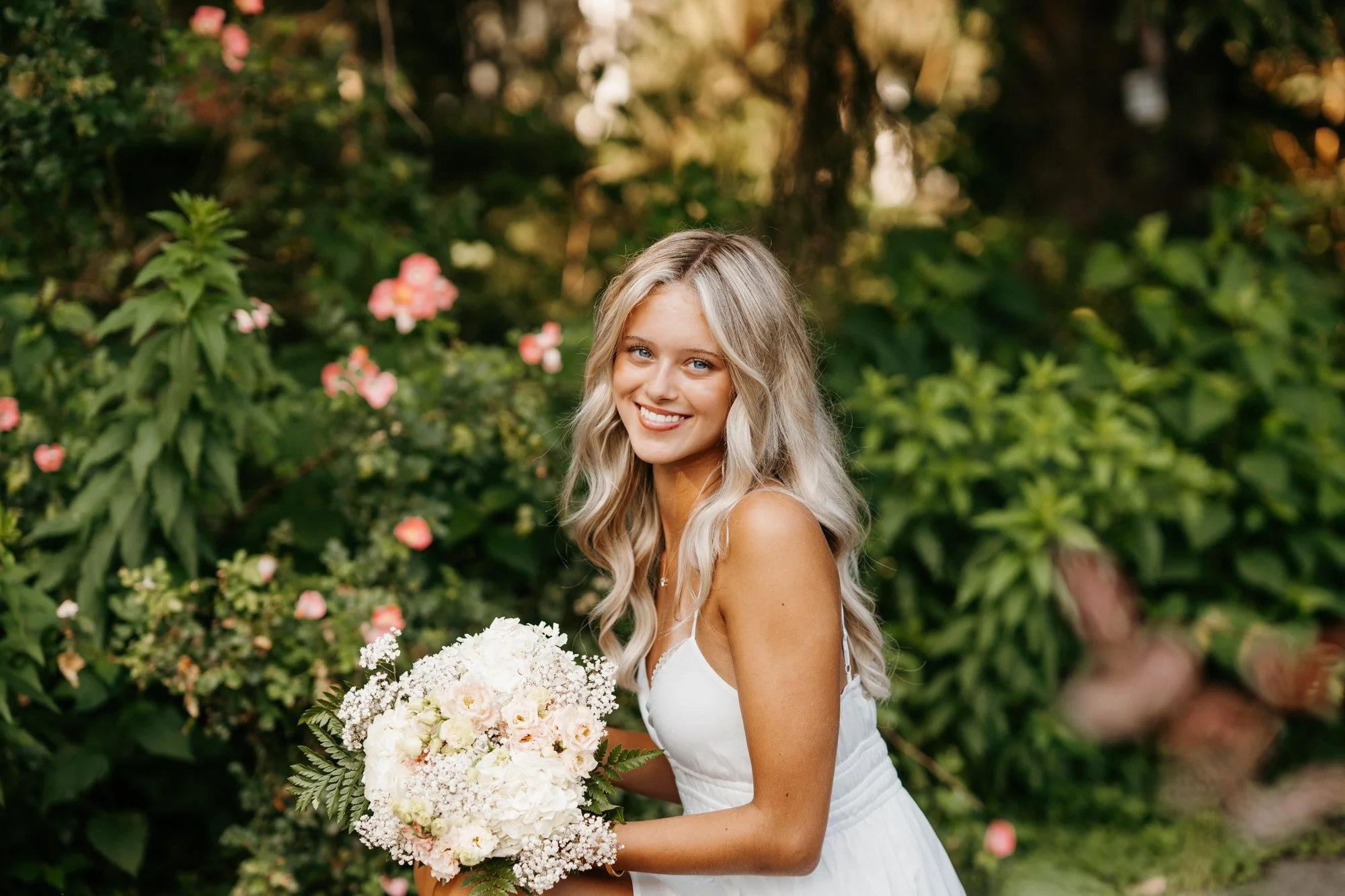 A young woman with long blonde hair, smiling, holding a bouquet of white and pale pink flowers, standing outdoors among green foliage and pink flowers.
