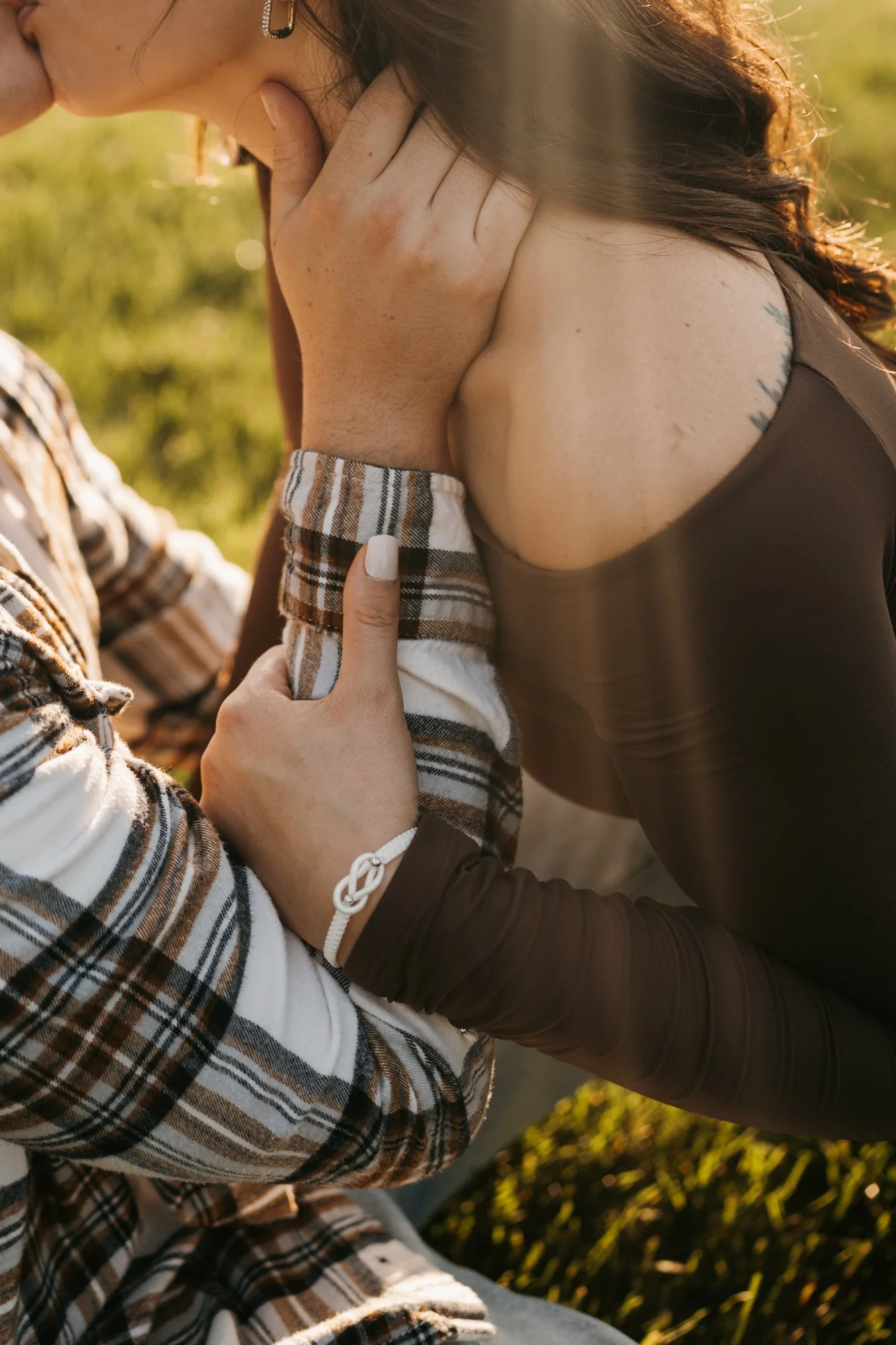 A close-up of two people embracing outdoors during golden hour, with one person's hand gently holding the other's shoulder. The person on the right has dark hair, a tattoo on their shoulder, and is wearing a black top, while the person on the left ha