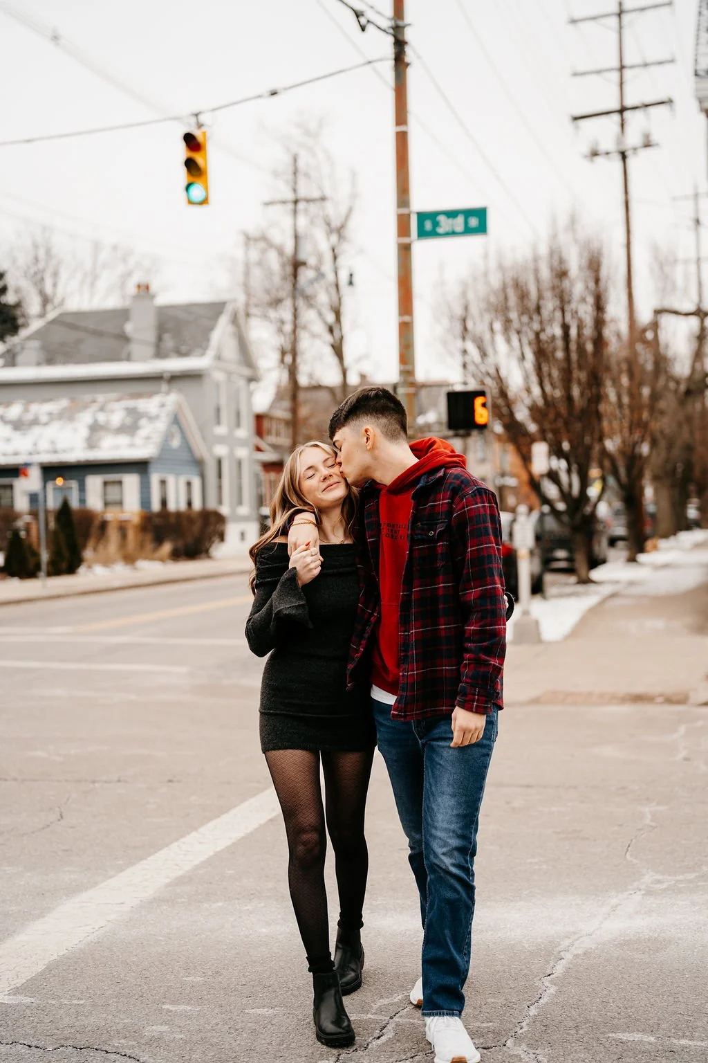 A young couple walking on a street, sharing a kiss, with snow on the ground and houses in the background.