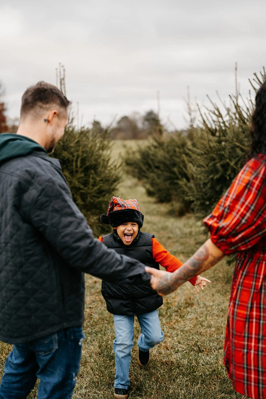 Three people, a man, a woman, and a young boy, holding hands and laughing outdoors in a Christmas tree farm with trees and cloudy sky in the background.