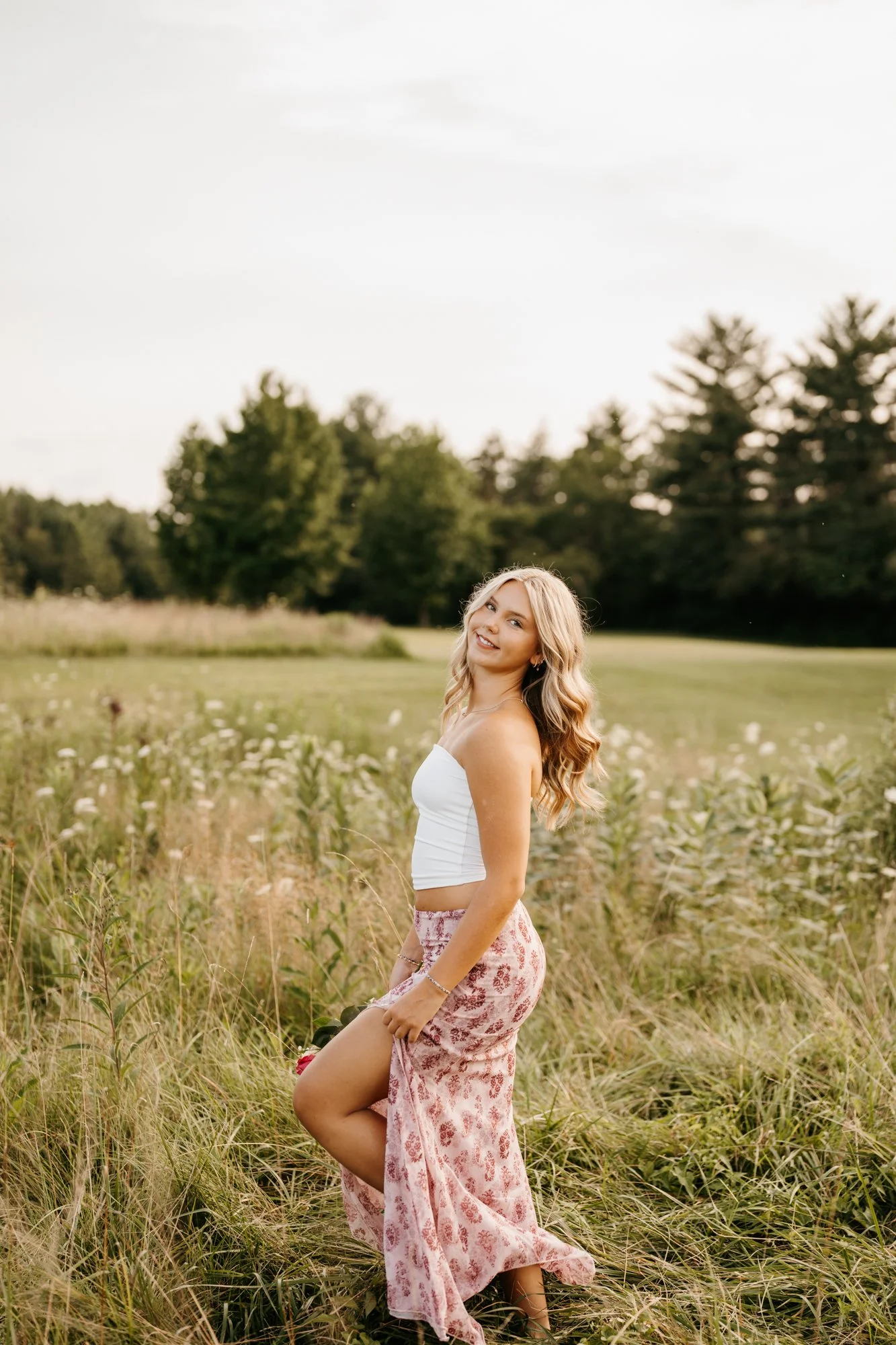 Young woman with long blonde hair in a white tank top and pink patterned skirt standing in a grassy field with trees in the background, smiling at the camera.