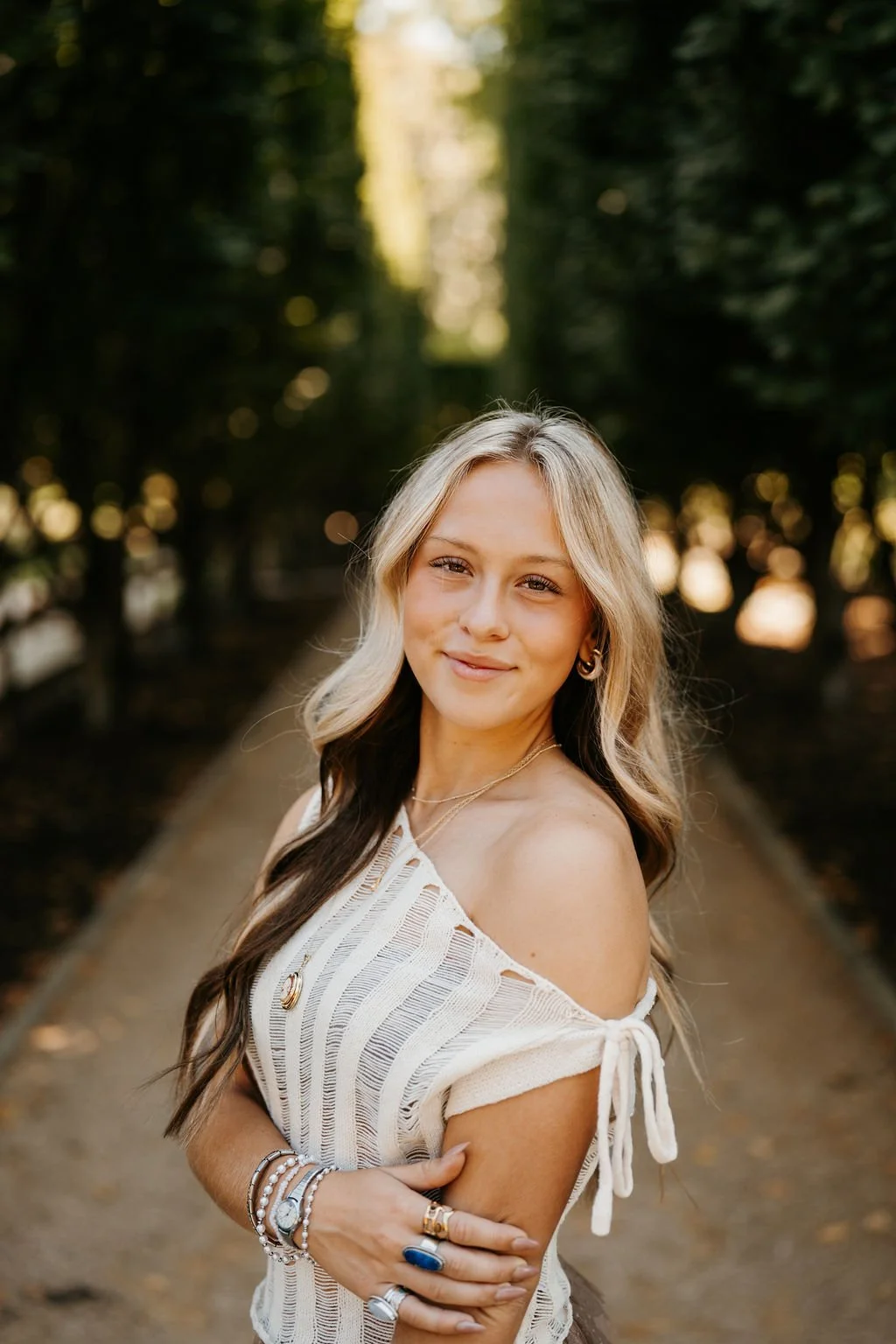 A young woman with wavy blonde and brown hair, wearing a white off-shoulder shirt, jewelry, and rings, smiling in a wooded outdoor setting with sunlight filtering through trees.