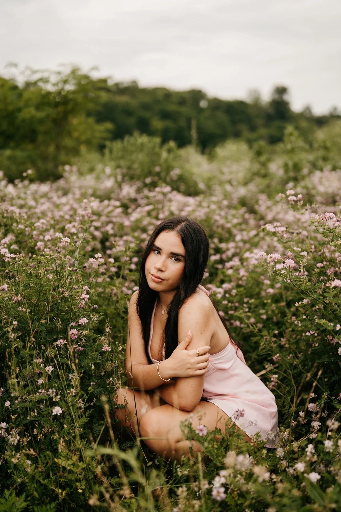 A young woman with long dark hair and a pink dress sitting in a field of pink flowers with green trees and overcast sky in the background.