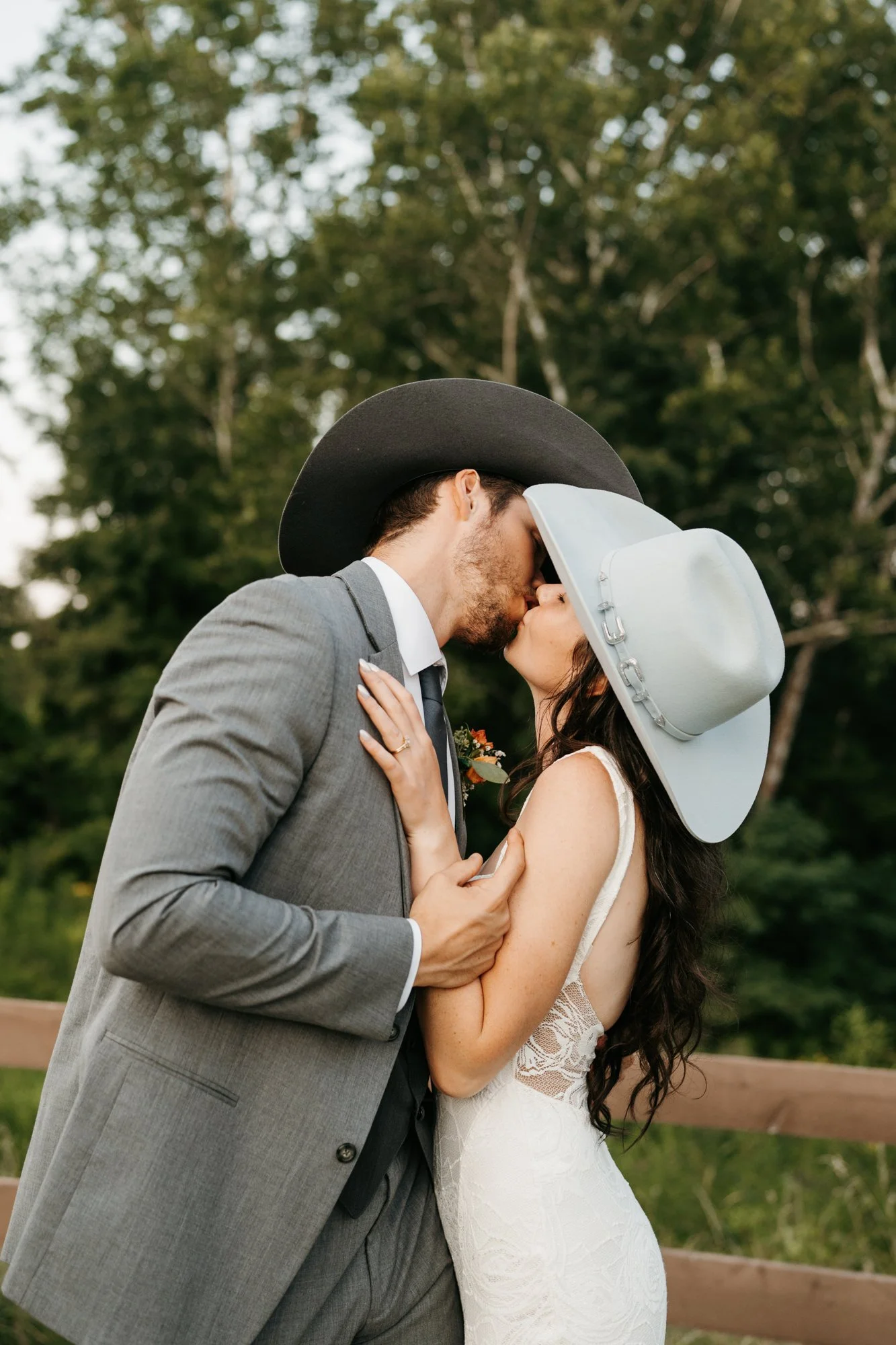 A couple, dressed in wedding attire, sharing a kiss outdoors. The man is wearing a gray suit with a black cowboy hat, and the woman is wearing a white lace wedding dress with a large white hat.