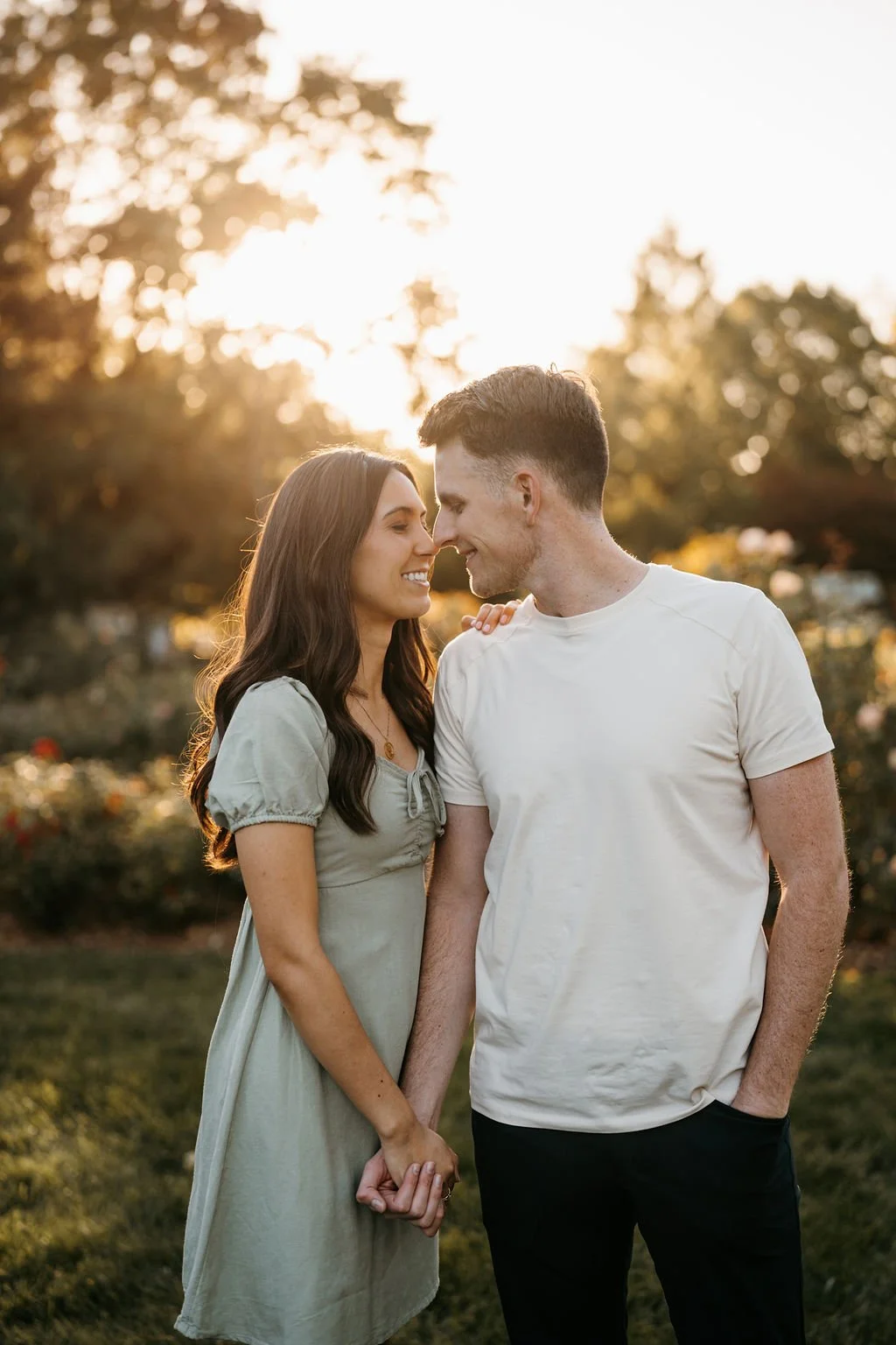 A young couple standing close together in a park at sunset, holding hands, smiling, with their foreheads touching.