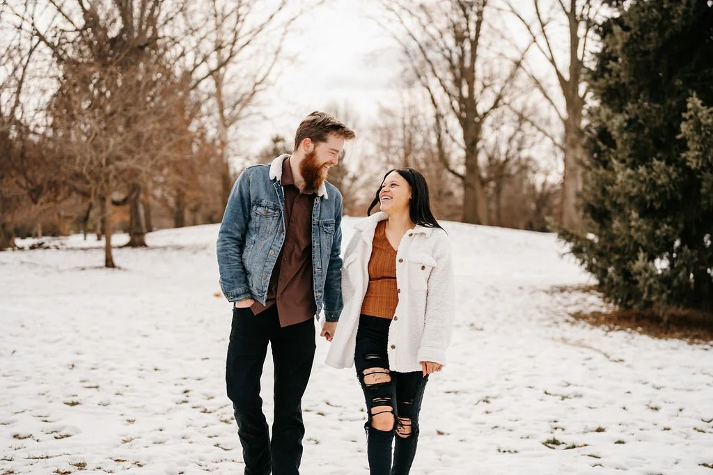 A man and woman walk together in a snowy park, smiling and laughing, holding hands.