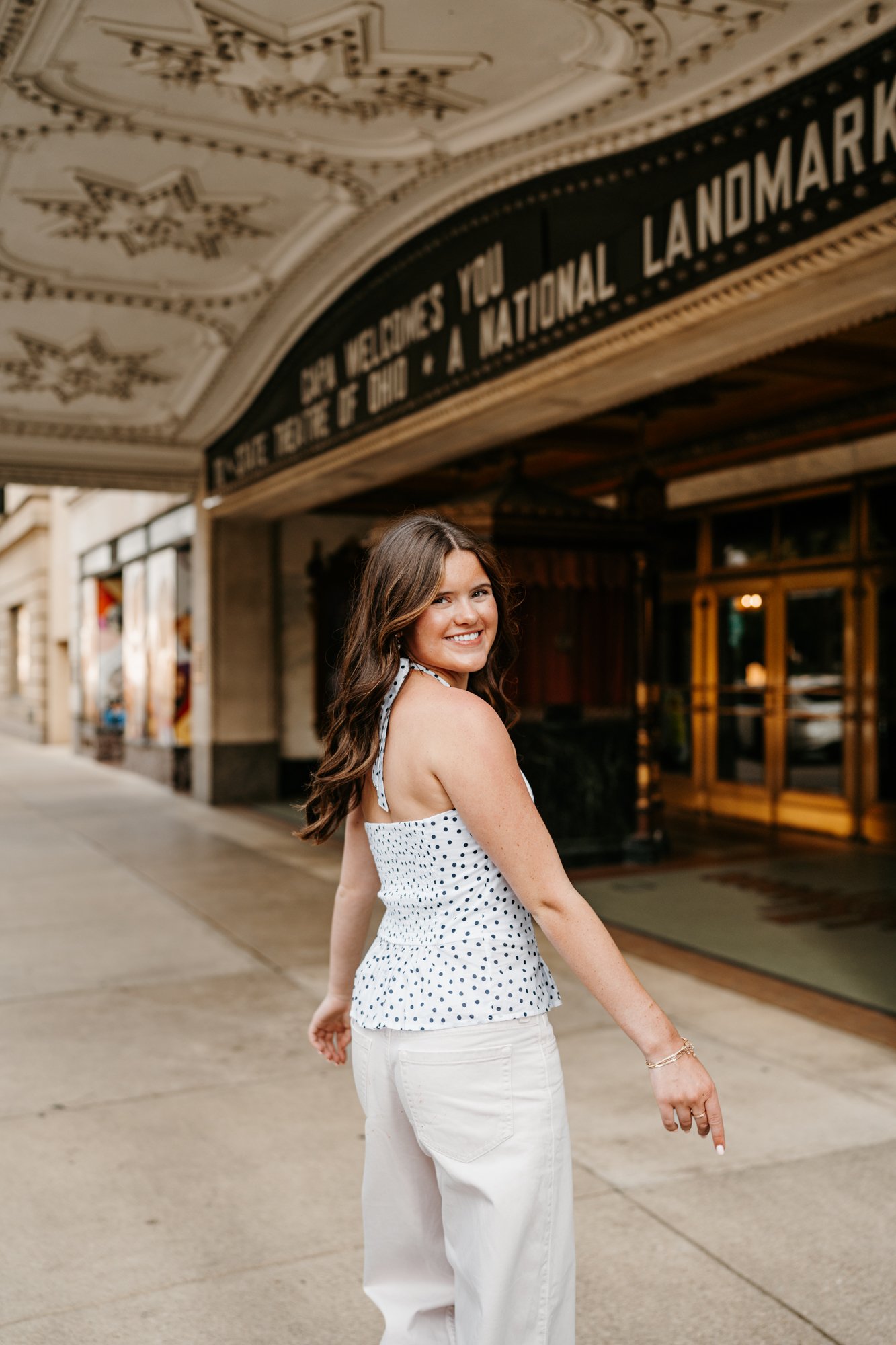 Young woman with long brown hair smiling and looking back, standing on a city sidewalk in front of a theater with a marquee, wearing a white polka dot top and white pants.