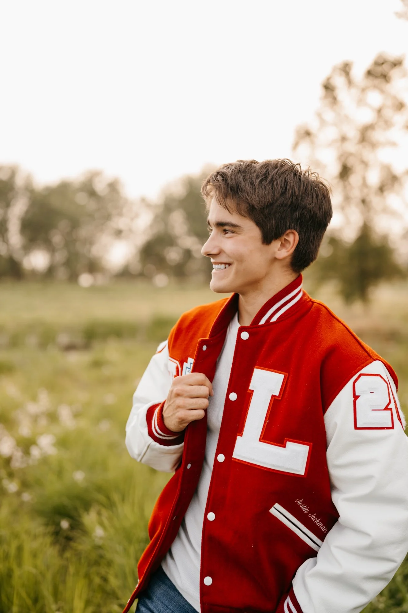 Young man with brown hair smiling, wearing a red and white varsity jacket with the letter L and number 2, standing outdoors in a grassy field with trees in the background.