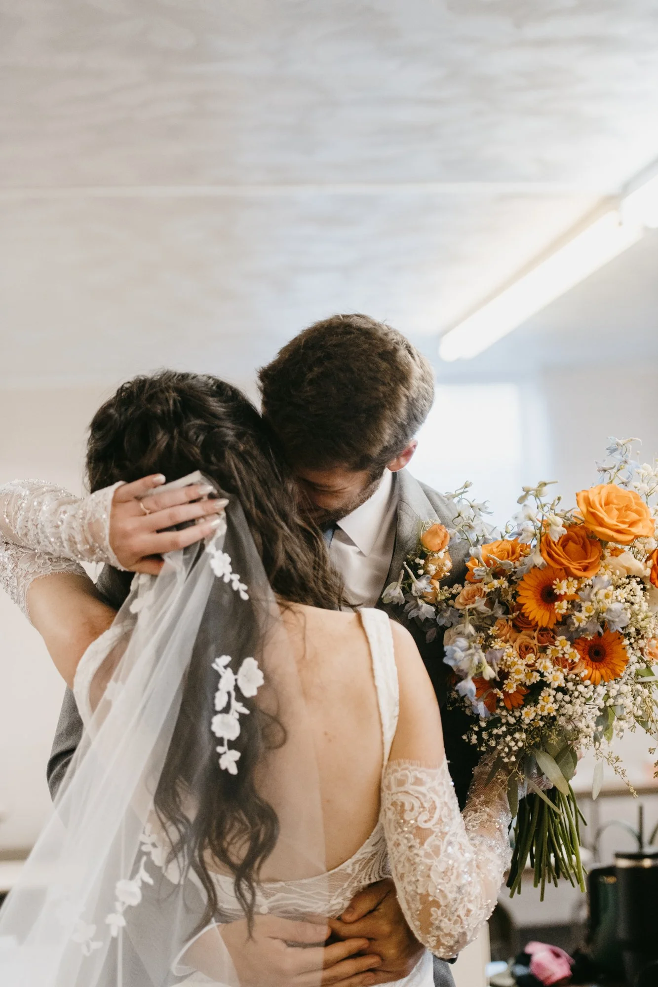 A bride and groom hug, with the groom holding a large bouquet of orange, yellow, and white flowers.