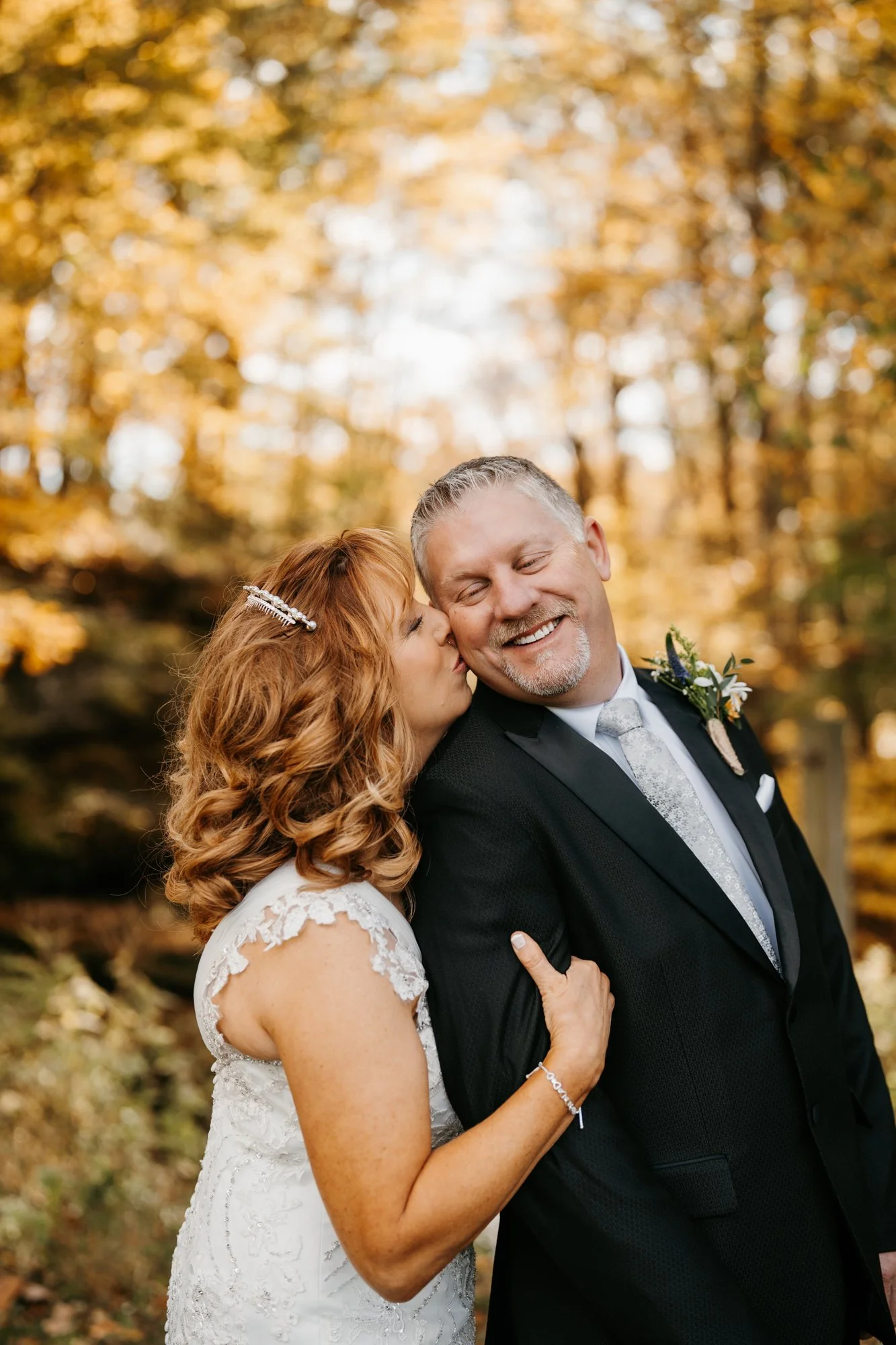 A bride with red hair in a white lace wedding dress kisses a smiling groom on the cheek, outdoors among autumn trees.