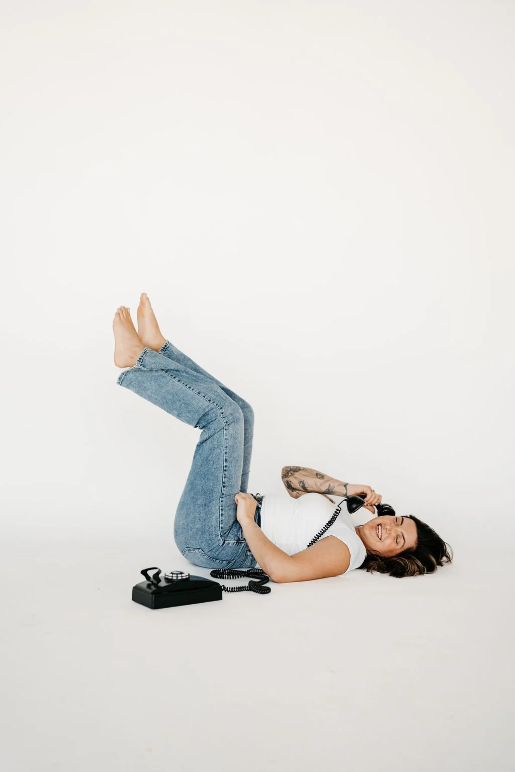 Woman lying on the floor, smiling, holding a vintage telephone receiver to her ear, with a rotary dial phone on the ground nearby.