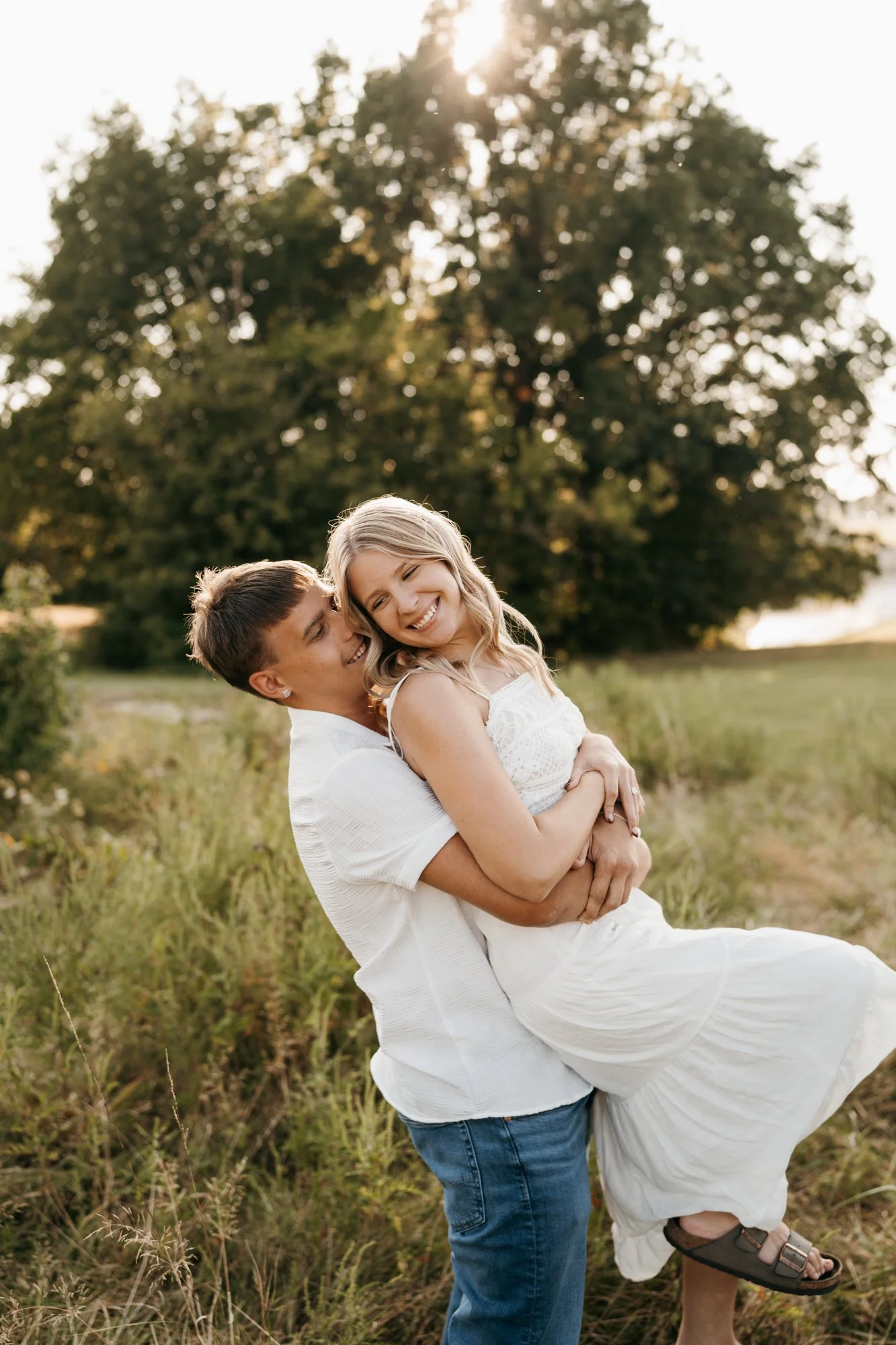 A young couple smiling and laughing outdoors, with the man holding the woman in his arms in a field with tall grass and a large tree in the background during sunset.