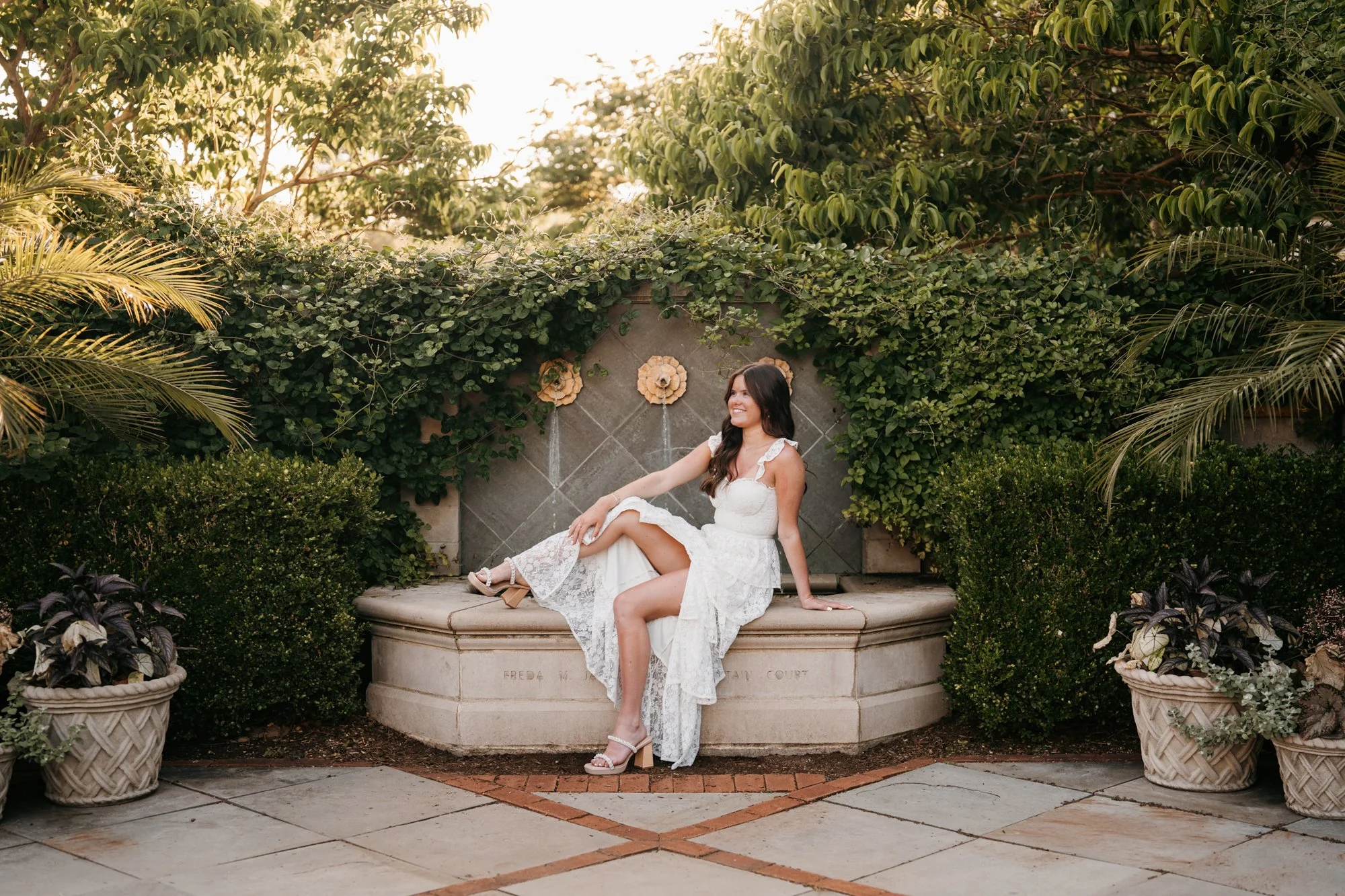 A woman in a white lace dress sitting on a stone fountain in a garden, surrounded by lush green plants and potted foliage, smiling with one leg crossed over the other.