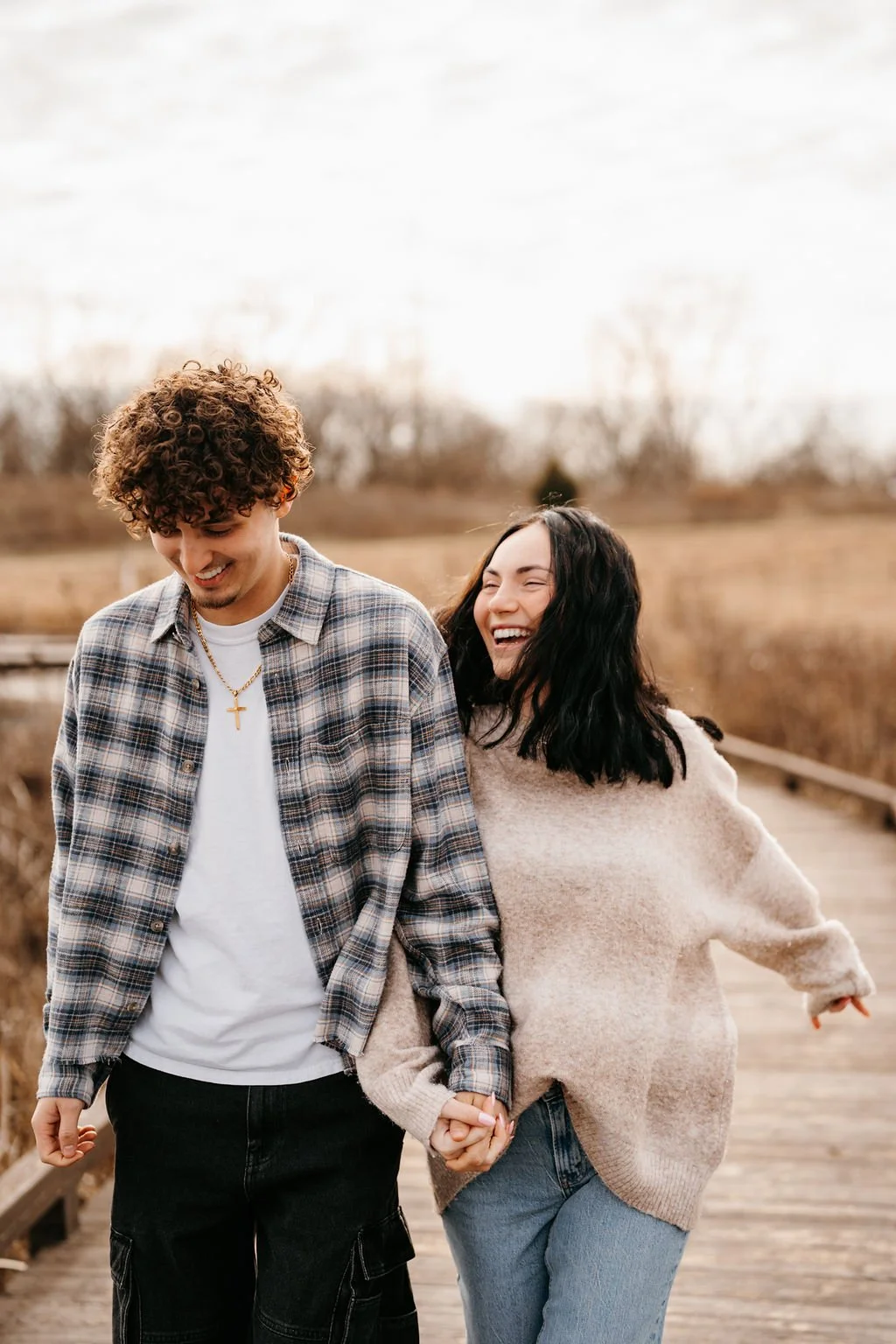 A happy couple walking outdoors on a wooden bridge, holding hands and smiling. The man has curly hair and is wearing a plaid shirt and a gold cross necklace. The woman has dark hair and is wearing a beige sweater and jeans.