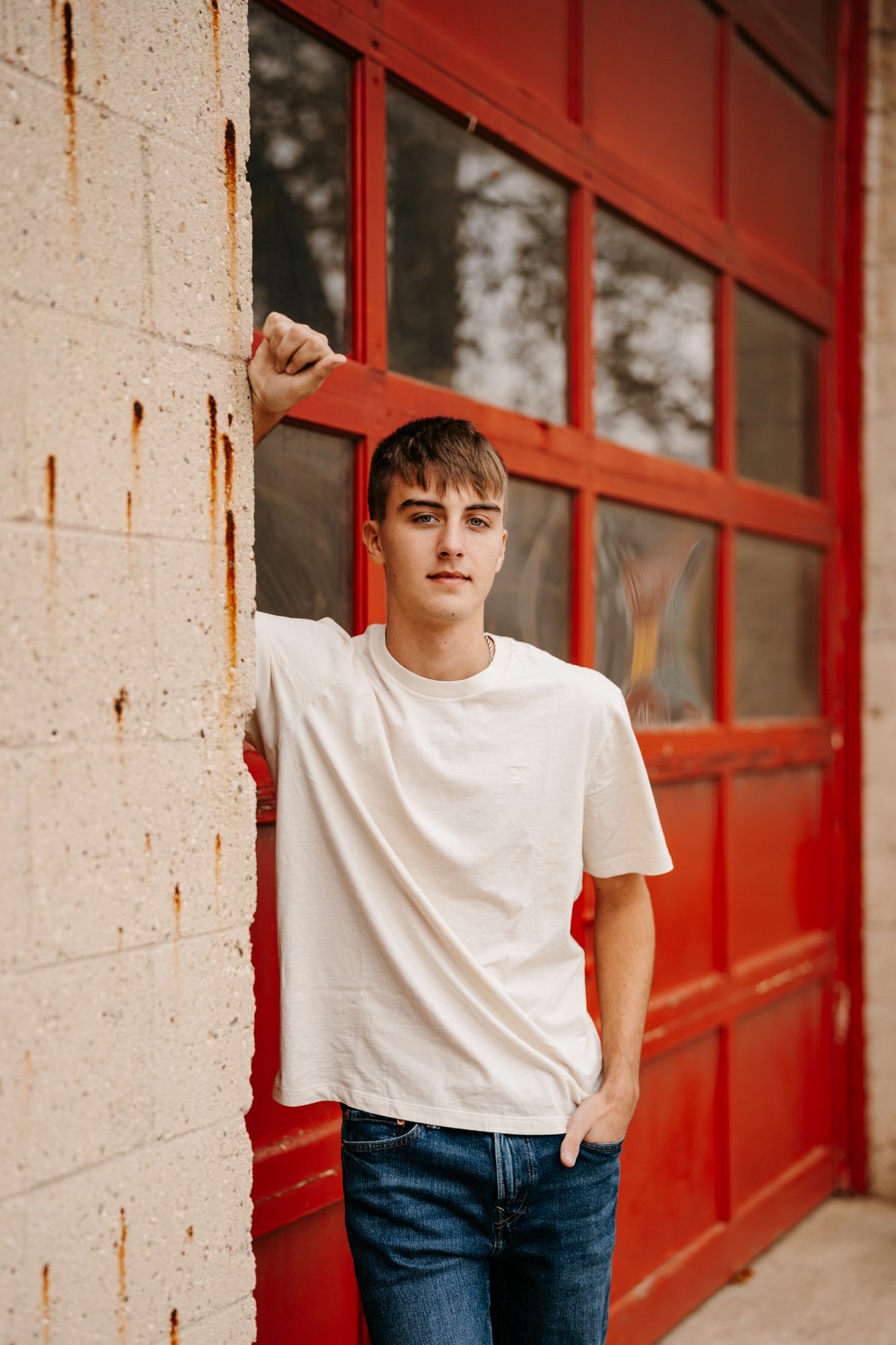 A young man in a white T-shirt and jeans standing outside next to a brick wall and a red garage door, leaning with his left arm raised and hand on the wall.