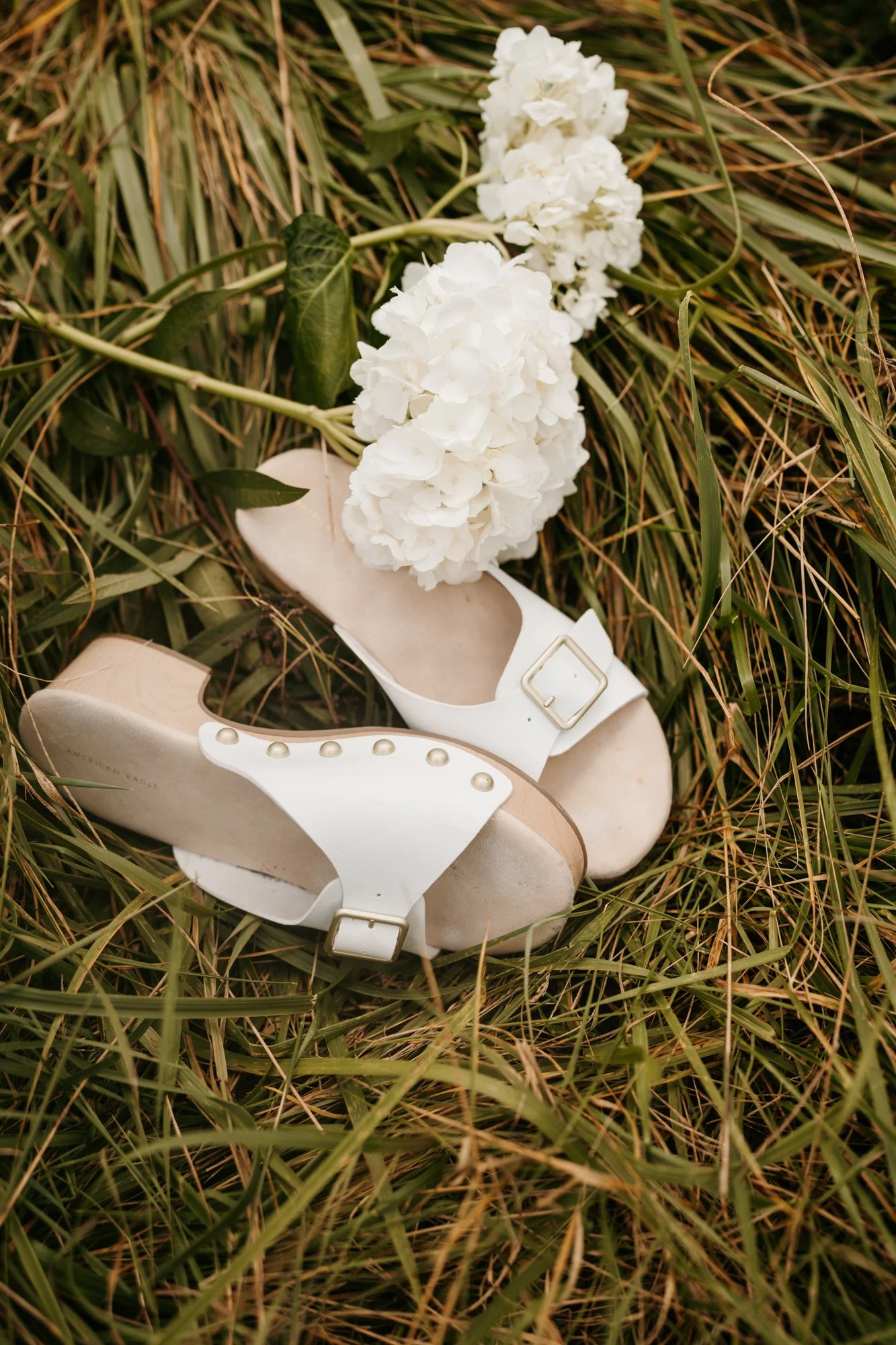 White heeled sandal with gold buckle, white flower bouquet, and green grass.