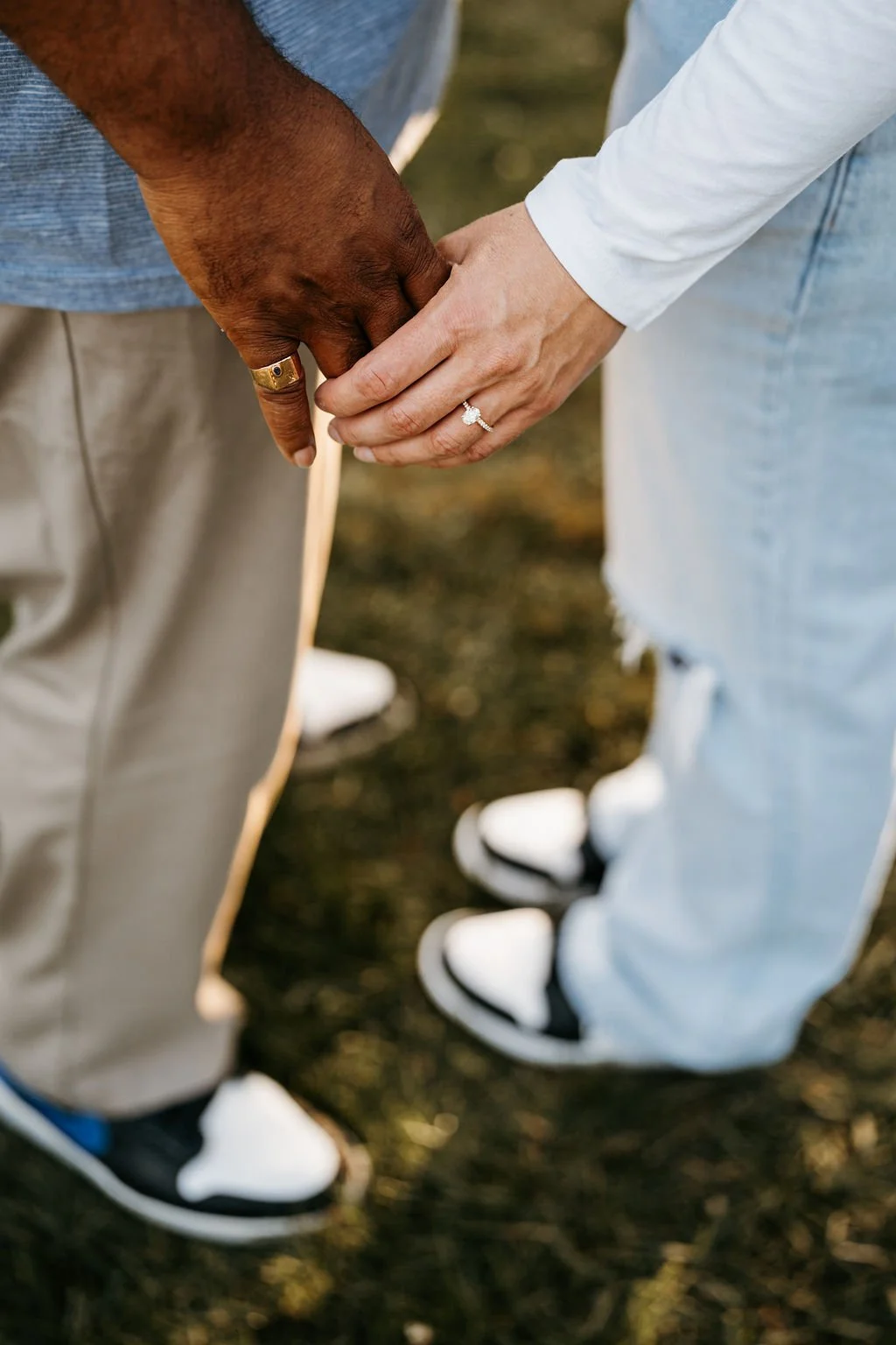 Close-up of two people holding hands, one with darker skin and a gold ring, the other with lighter skin and a diamond ring, outdoors on grass.