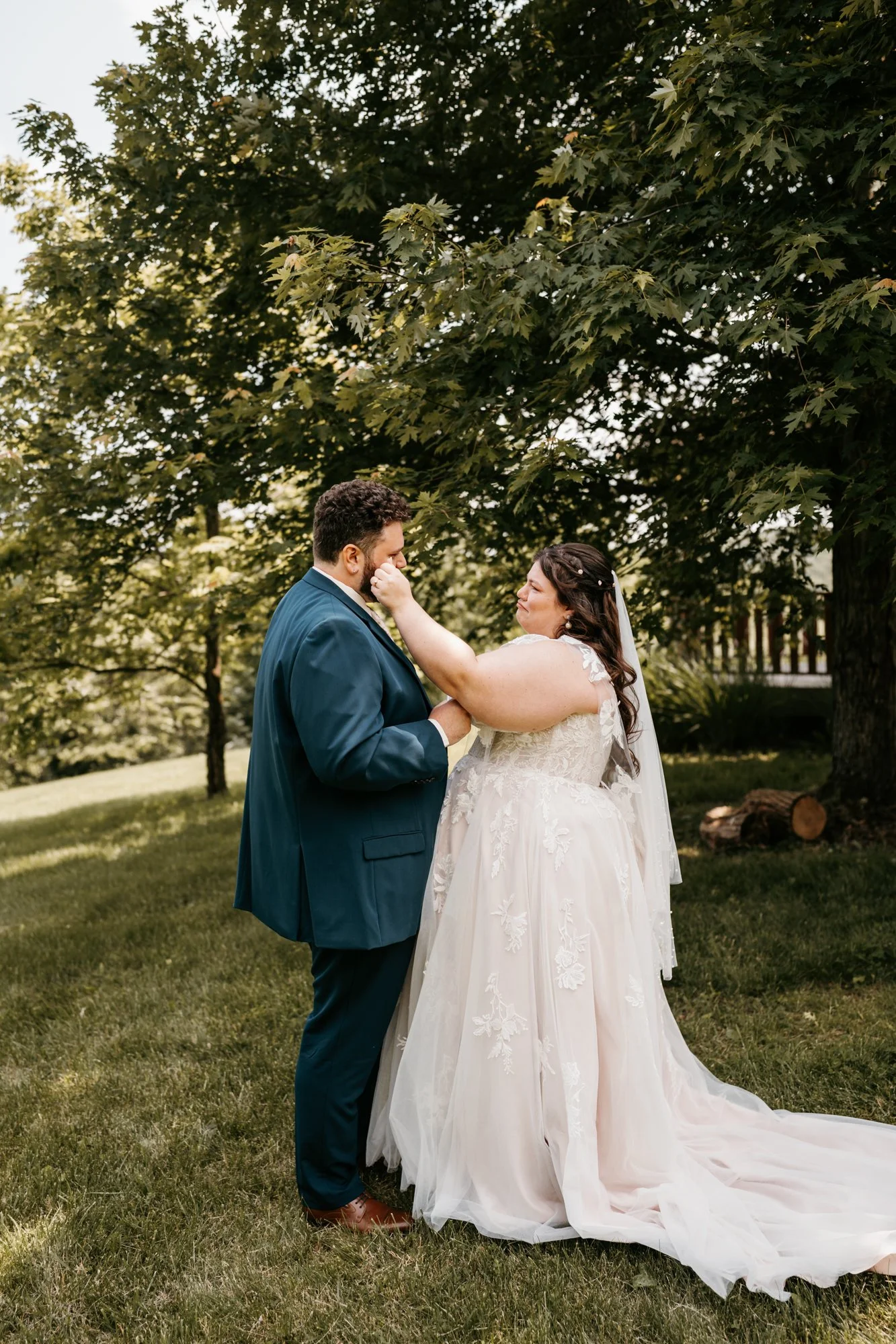 Bride and groom standing outdoors, with the bride touching the groom's face, under a large leafy tree.