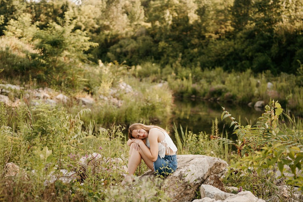A young woman sitting on a rock by a river, surrounded by lush greenery and trees during daylight.