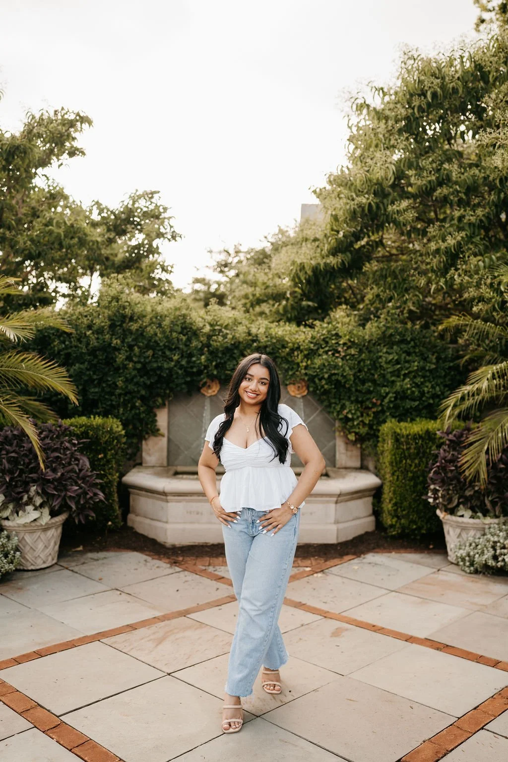 A smiling woman in a white top and light blue jeans standing outdoors in front of a garden backdrop with fountain and greenery.