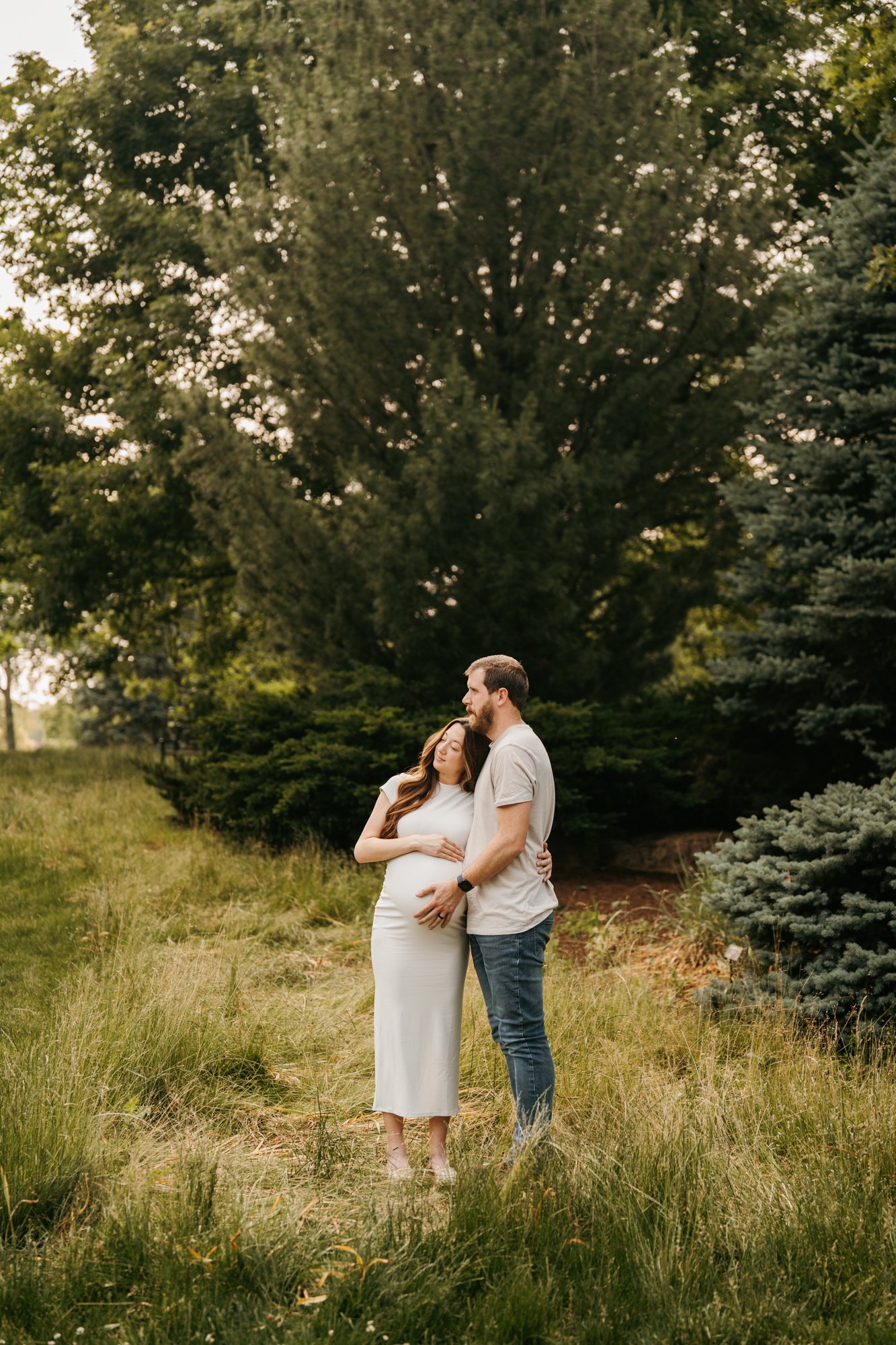 A pregnant woman and a man standing in a grassy outdoor setting with trees in the background, embracing lovingly.