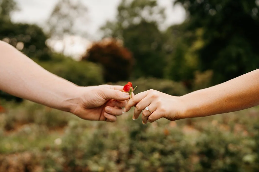 Two hands reaching out, one hand holding a small red flower, and the other hand with an engagement ring, outdoors with blurred trees in the background.