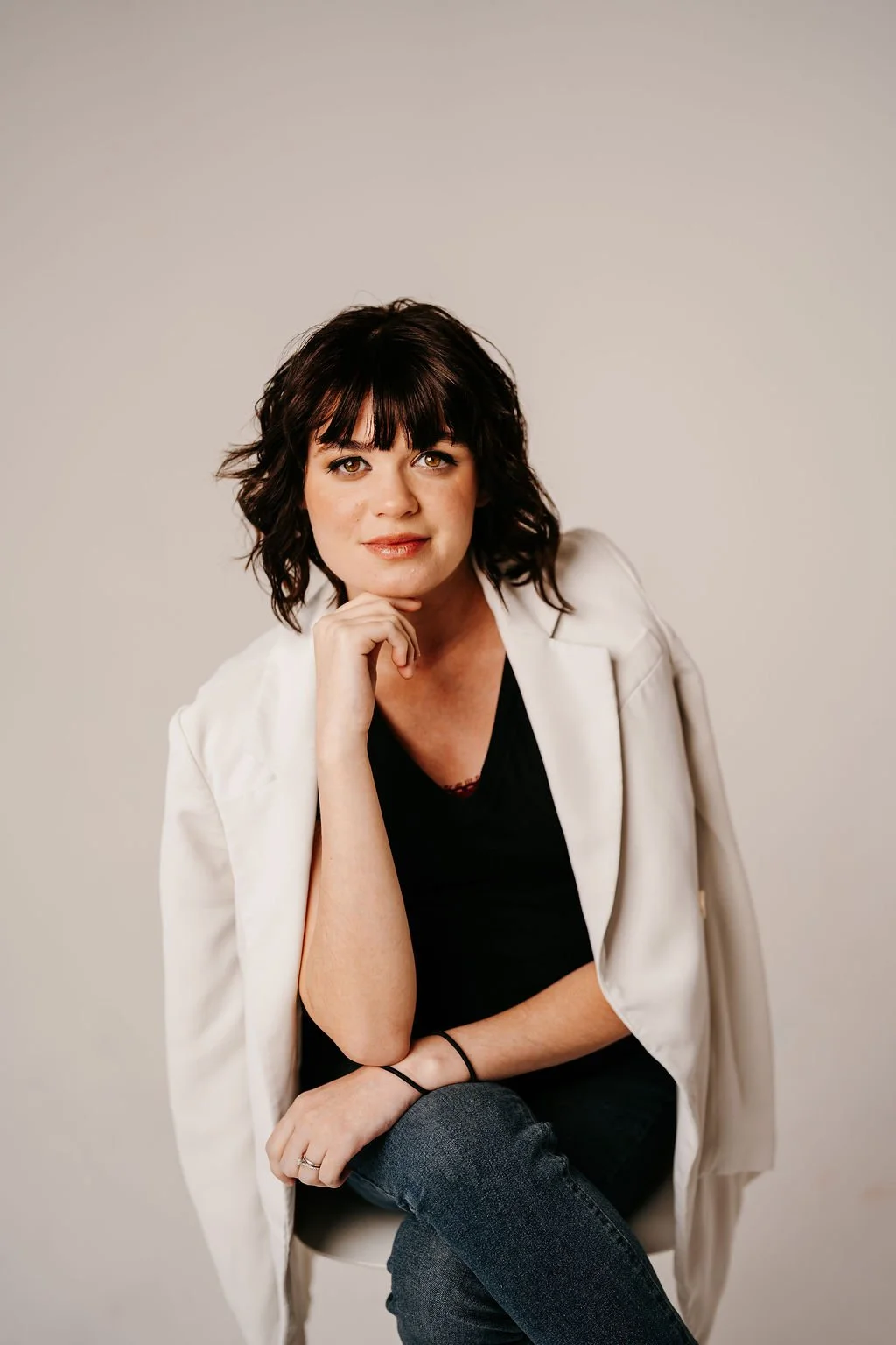 Portrait of a woman with dark, shoulder-length wavy hair, wearing a white blazer over a black top, sitting against a plain background.