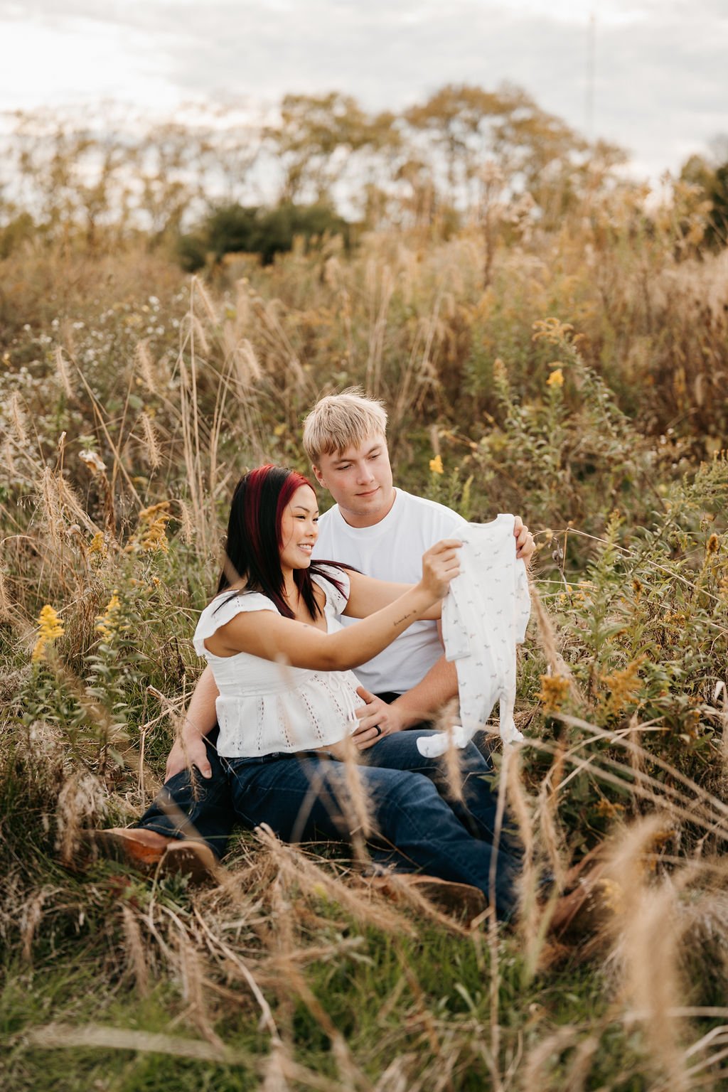 A couple sitting in a grassy field with dry and green plants during sunset, smiling and holding a baby onesie.
