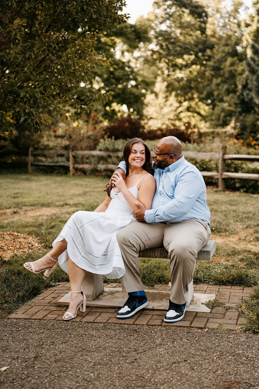 A smiling woman in a white sundress and beige high-heeled sandals sits on a bench next to a bearded man in a light blue shirt, beige pants, and sneakers, smiling and looking at each other in a park with trees and a wooden fence in the background.
