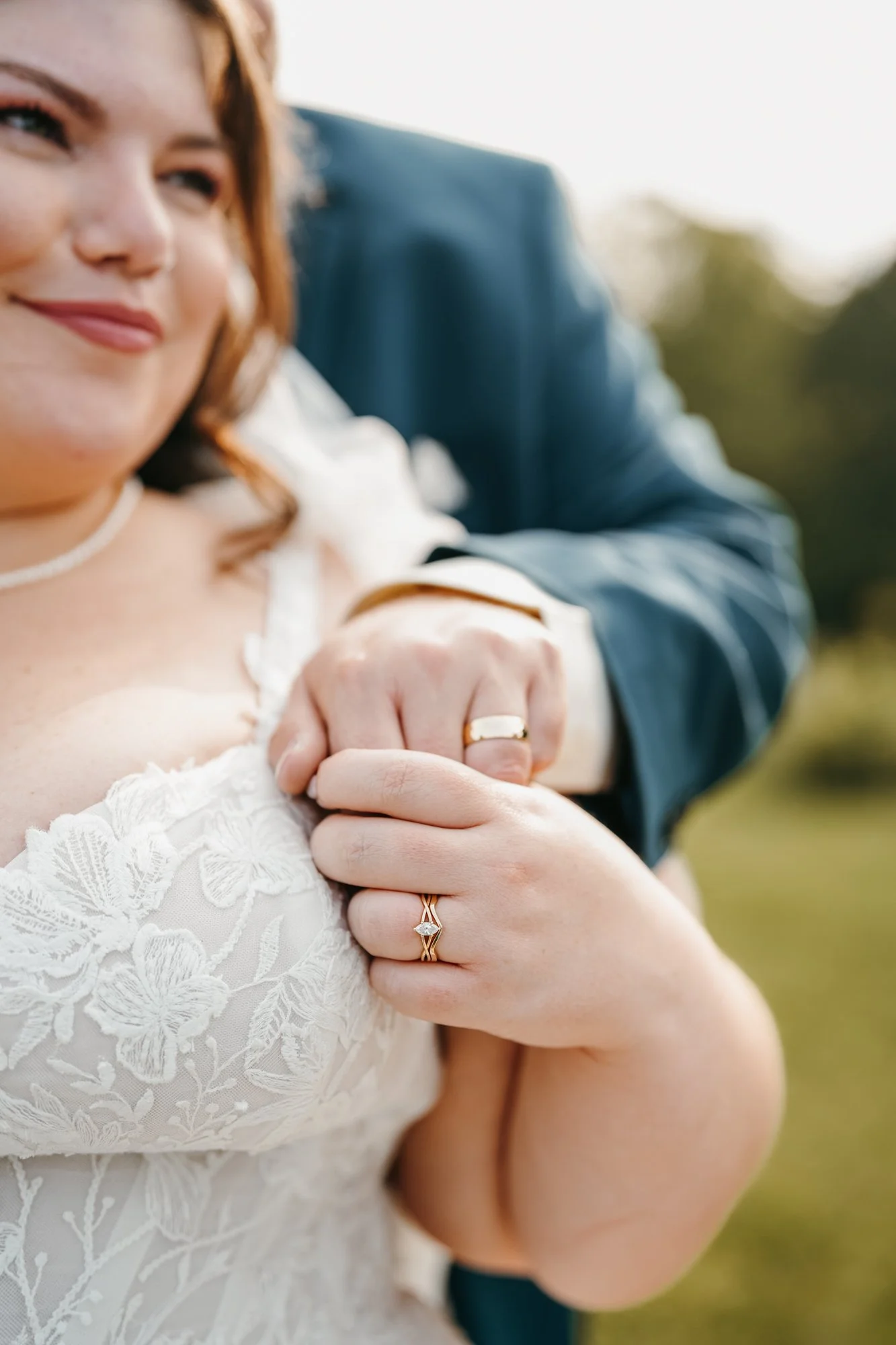 Close-up of a bride and groom holding hands, showcasing wedding rings, with a blurred green background.
