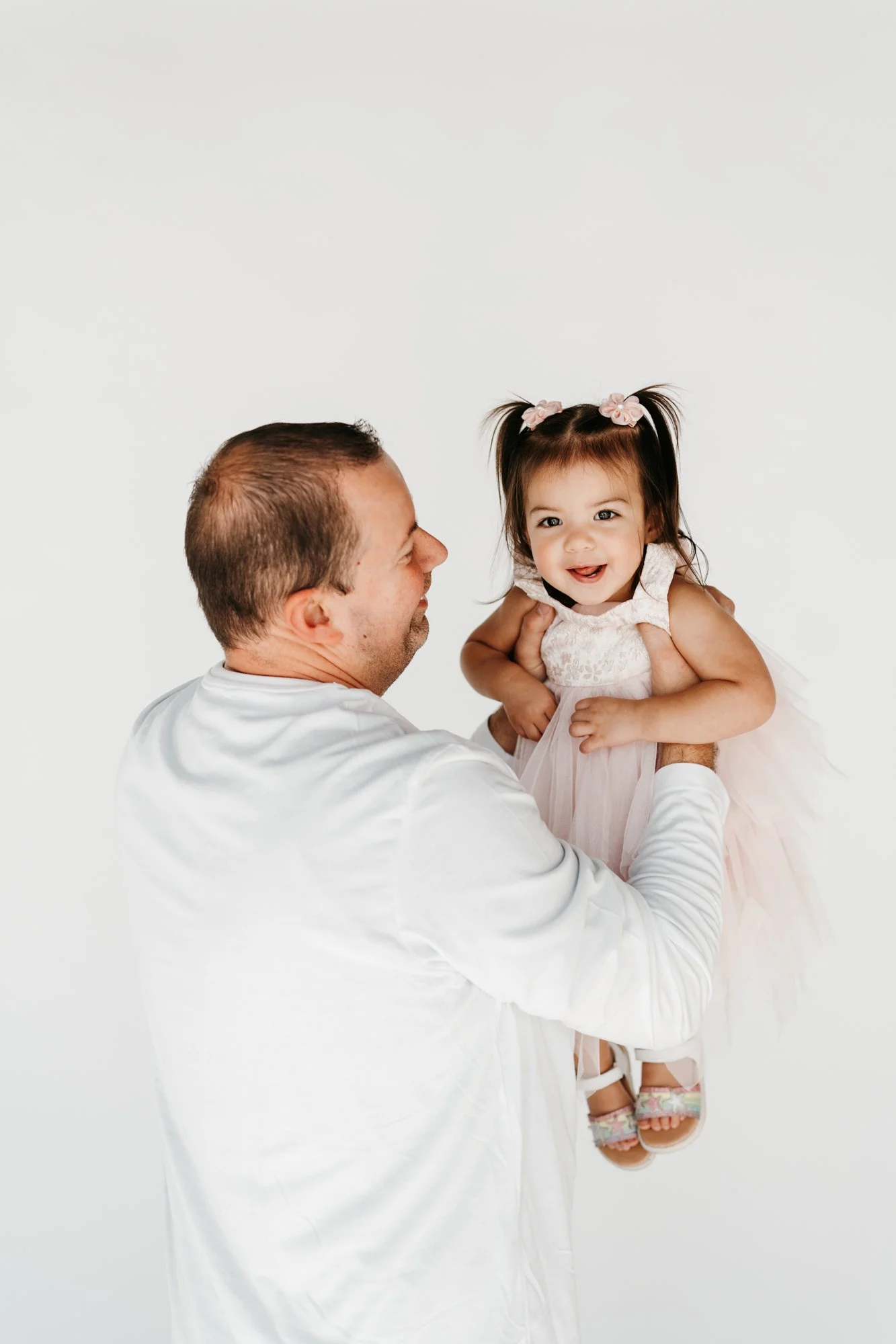 A man holding up a smiling young girl with pigtails and floral hair accessories, against a plain white background.