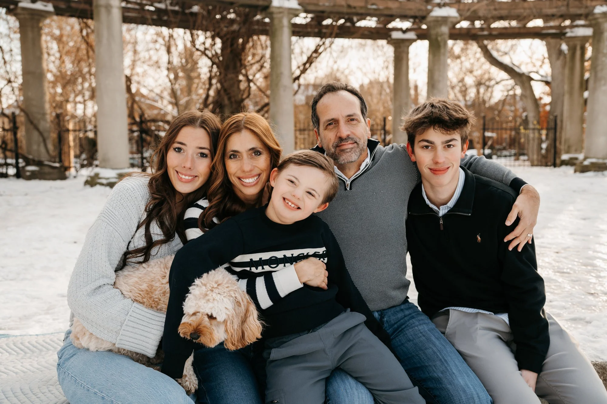 A family of five, including three children, an adult woman, and an adult man, sitting outdoors in a snowy park with leafless trees and pillars in the background, smiling at the camera, with a dog on the woman's lap.