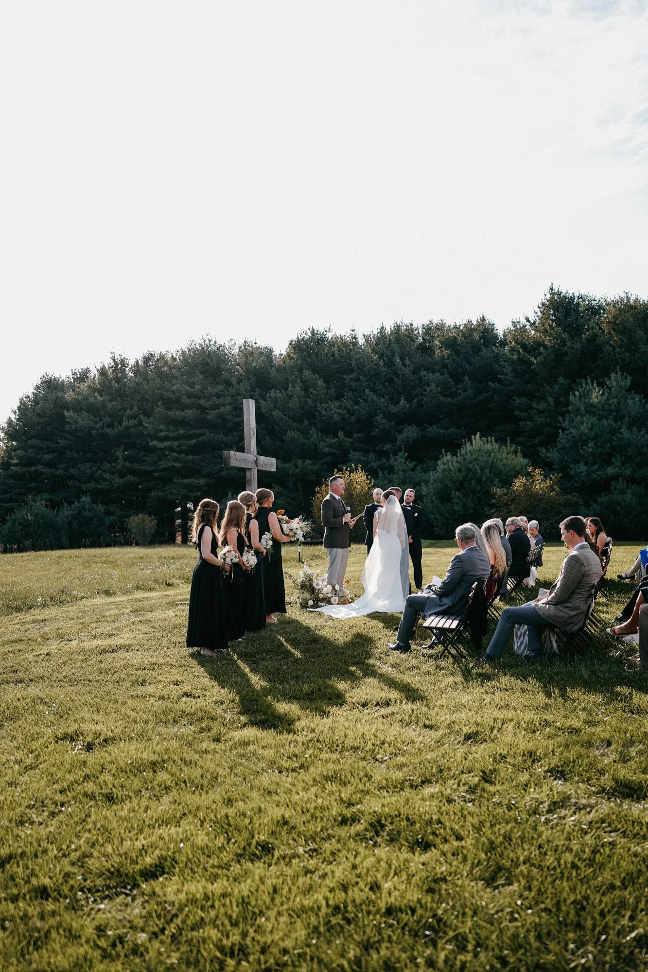 A wedding ceremony outdoors on green grass, with the bride and groom facing each other, standing near a large wooden cross, surrounded by wedding party and guests, with trees in the background, under a cloudy sky.