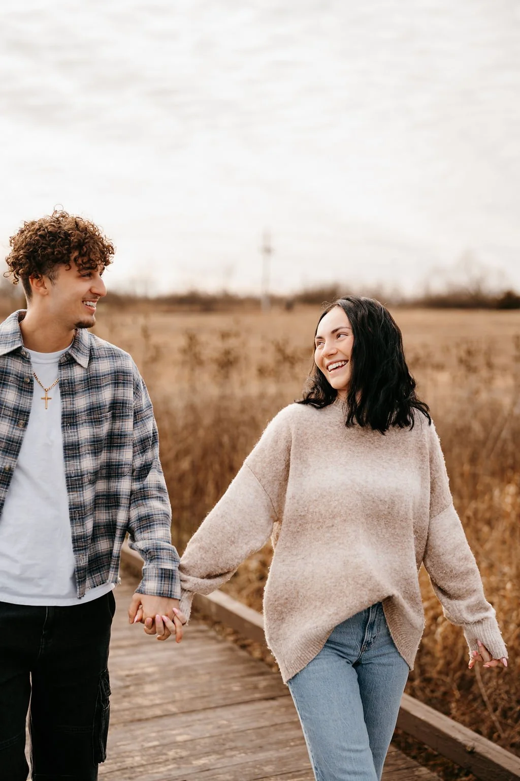 A young man and woman holding hands and smiling at each other while walking on a wooden path through a grassy field.
