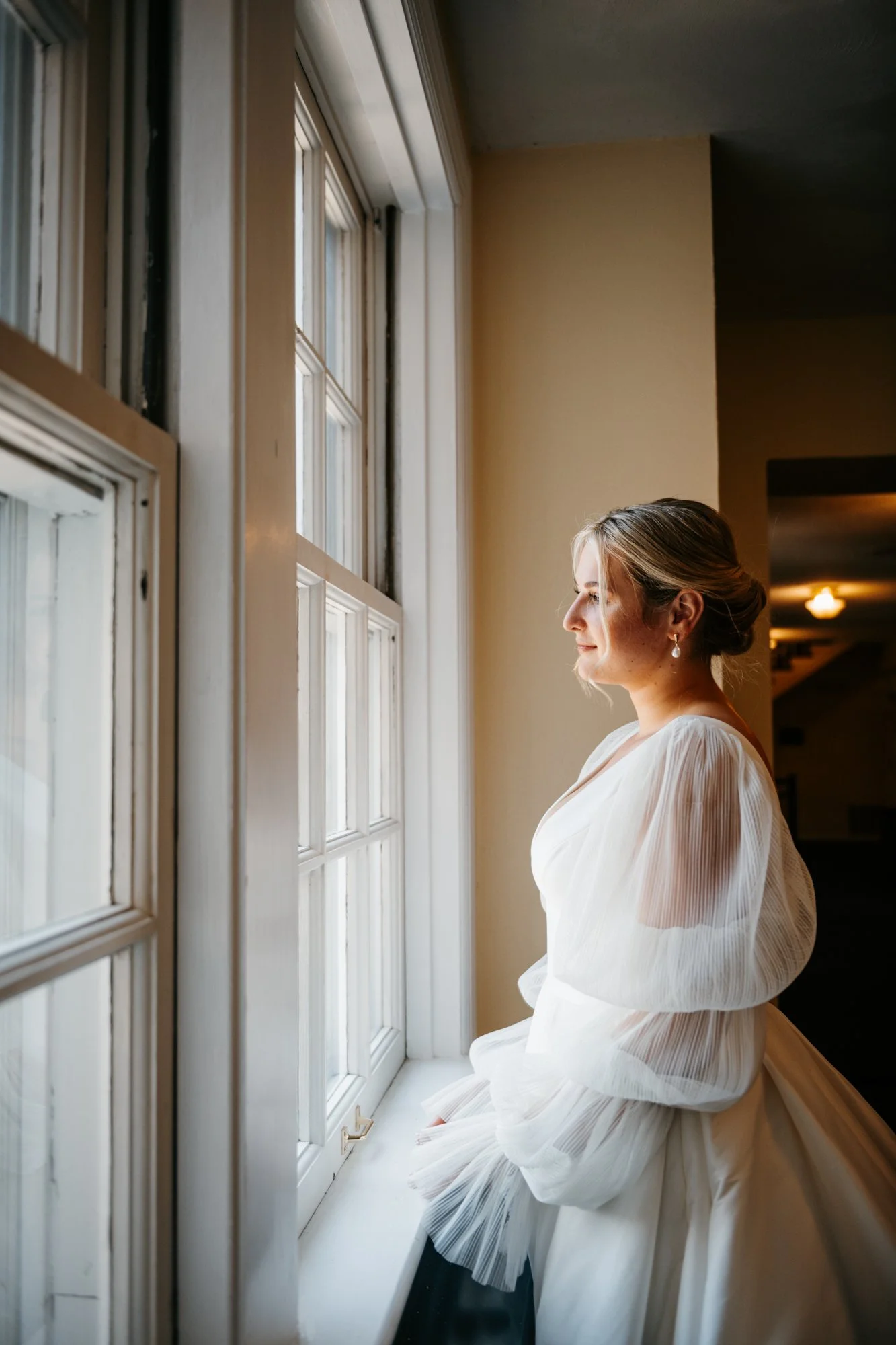 A woman with blonde hair styled in an updo, wearing a white dress with puffed sleeves, gazes out a window with divided panes in a softly lit room