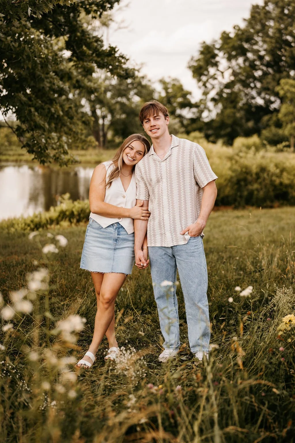 A young couple standing hand in hand outdoors near water, smiling, with trees and grass around them.
