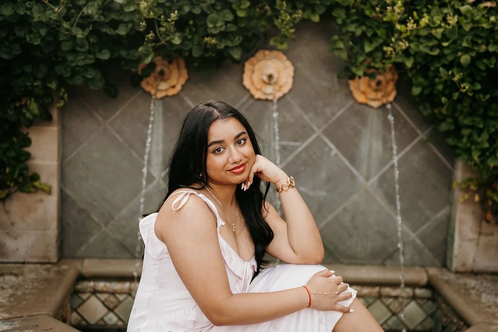 Young woman sitting outdoors, wearing a light pink dress, with dark hair and makeup, sitting in front of a tiled wall with decorative flowers and greenery.